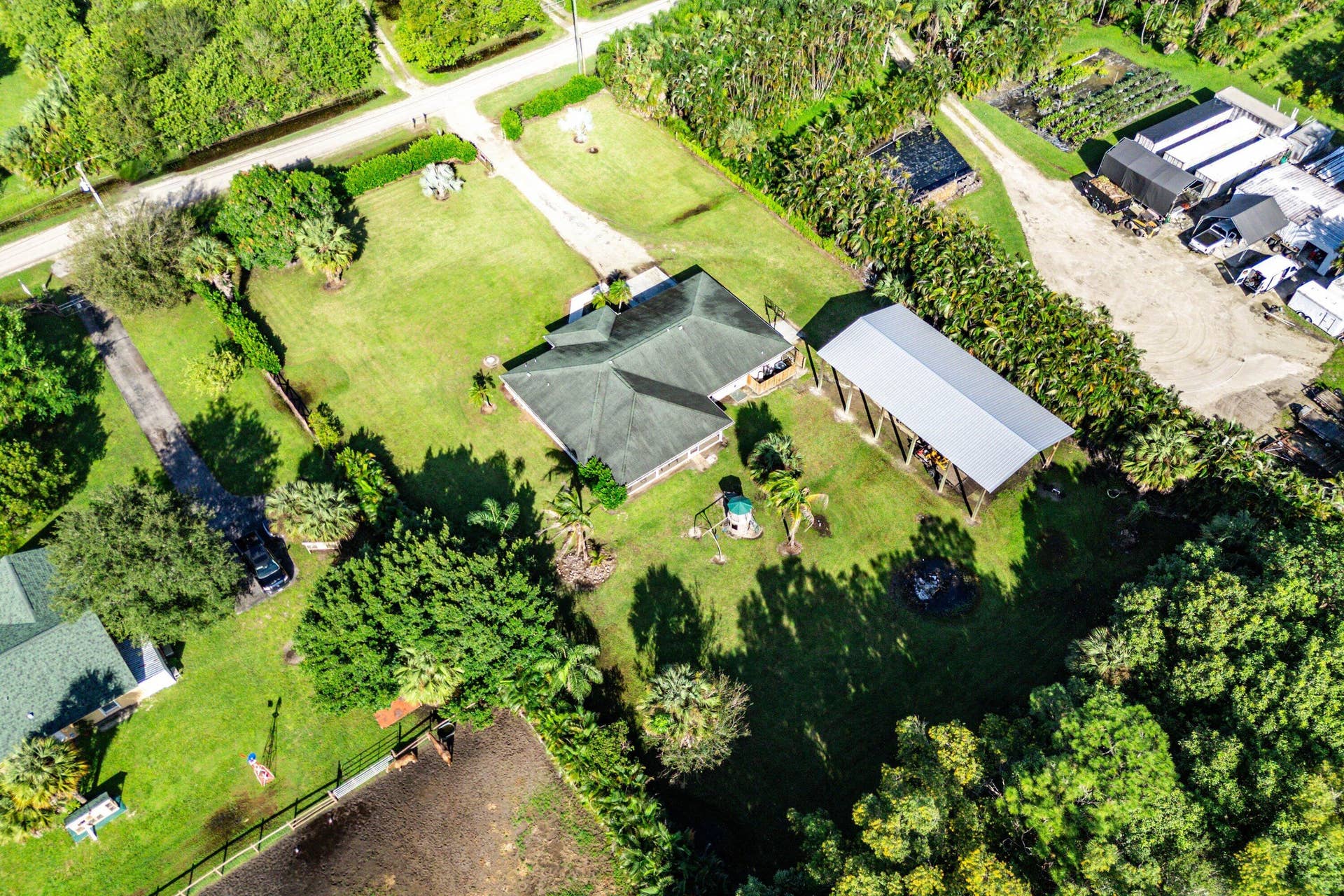 Aerial perspective of a sprawling property featuring a multi-hipped roofline, an expansive lawn, mature palm trees, a detached open-air metal-roof structure, and perimeter fencing