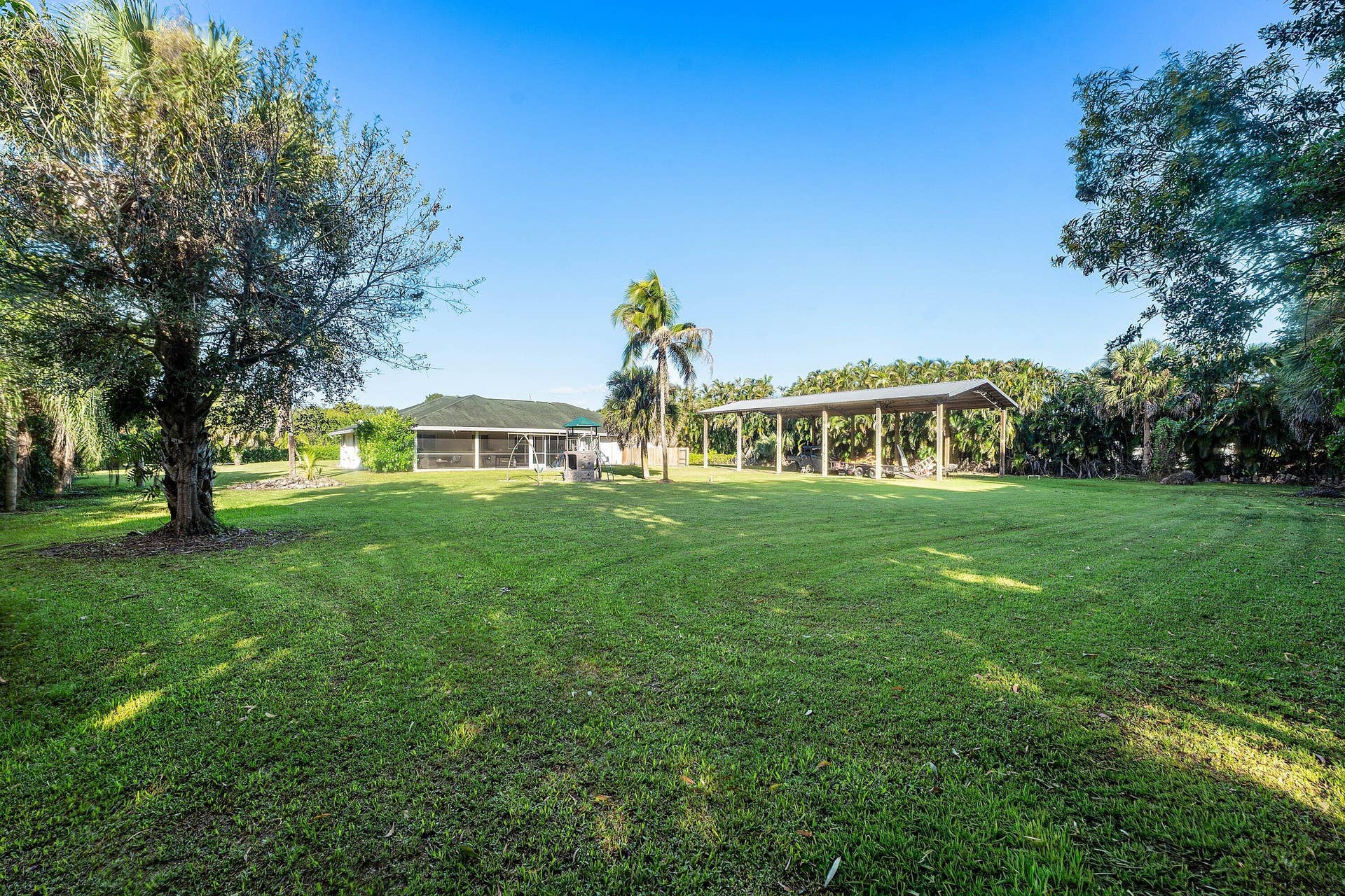 Expansive backyard featuring a sprawling lawn, mature trees, open-air metal carport, and screened rear patio