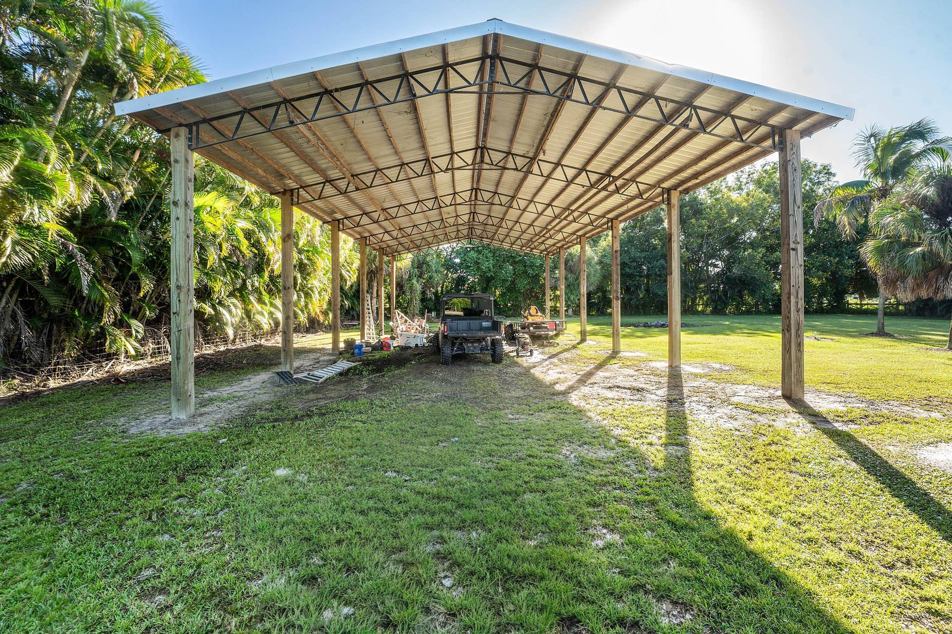 Open-air structure featuring a metal roof, steel truss framework, and heavy-duty timber support posts situated on an expansive grass lot