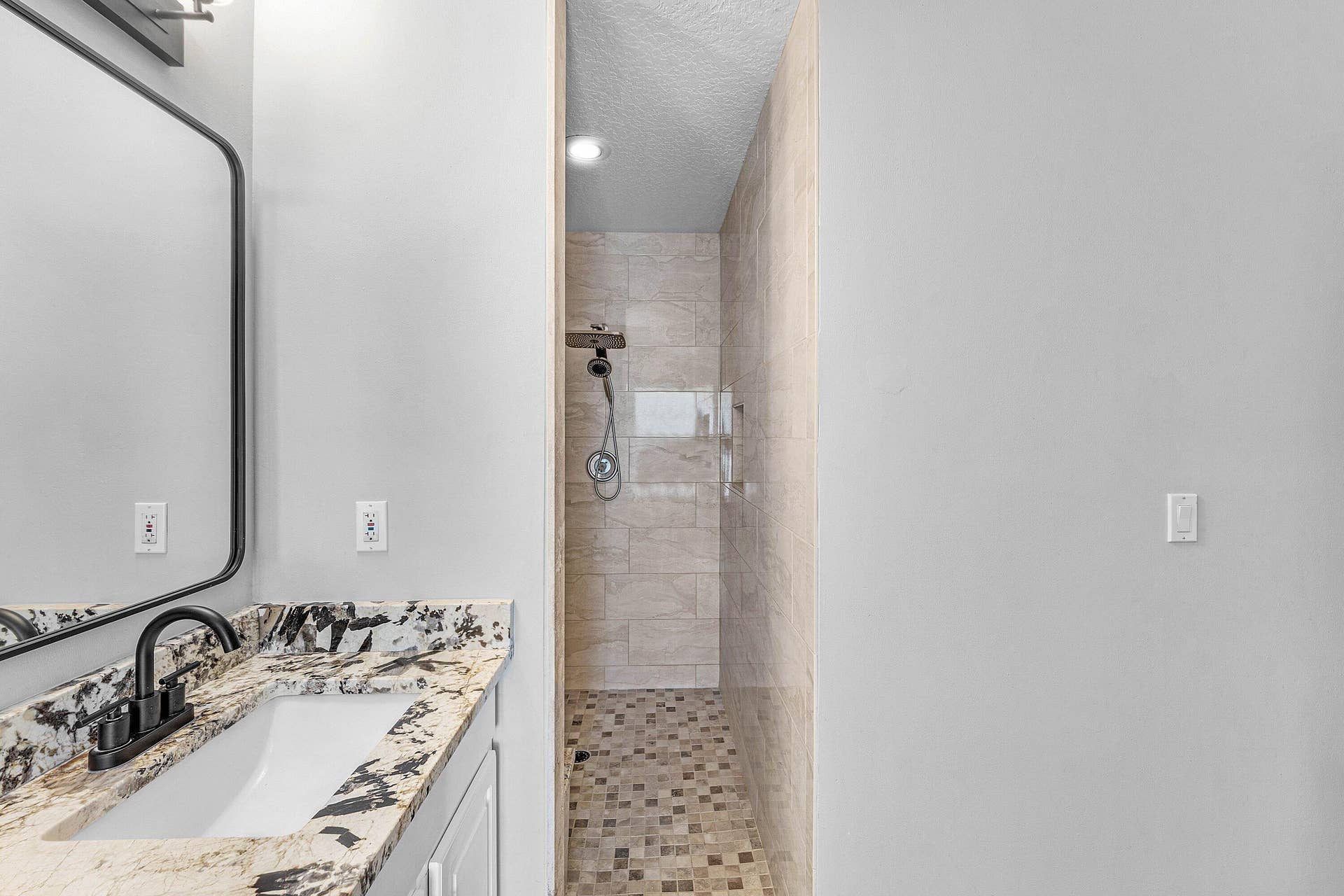 Vanity with stone countertop and undermount sink, matte black faucet, wall-mounted mirror, and walk-in shower with stone tile wall surfacing and mosaic tile flooring