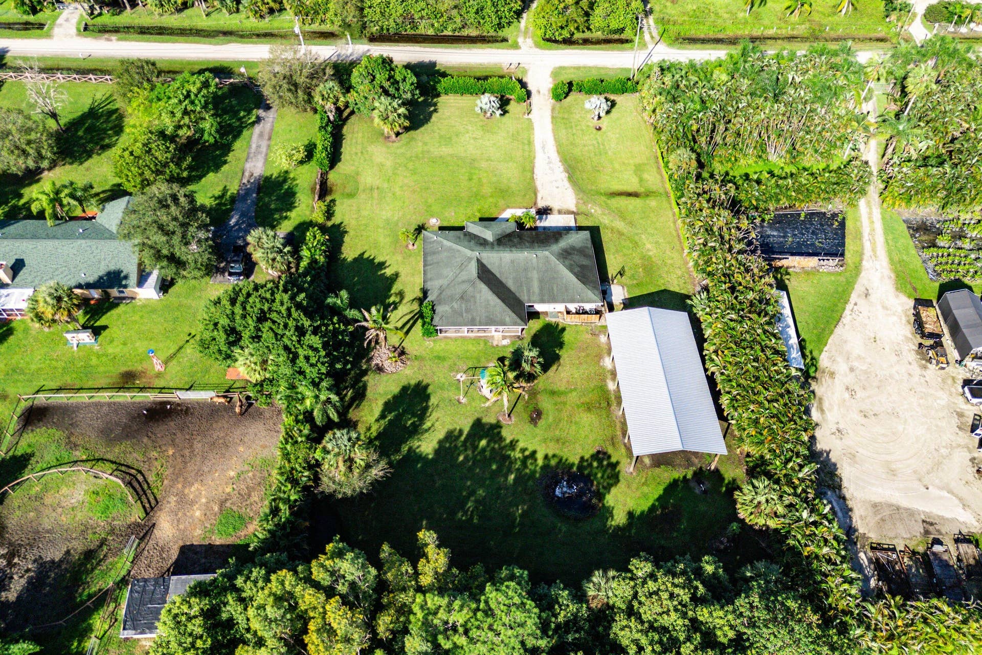 Aerial perspective of a sprawling lot featuring a detached metal-roof structure, expansive green space, mature landscaping, and a fenced enclosure