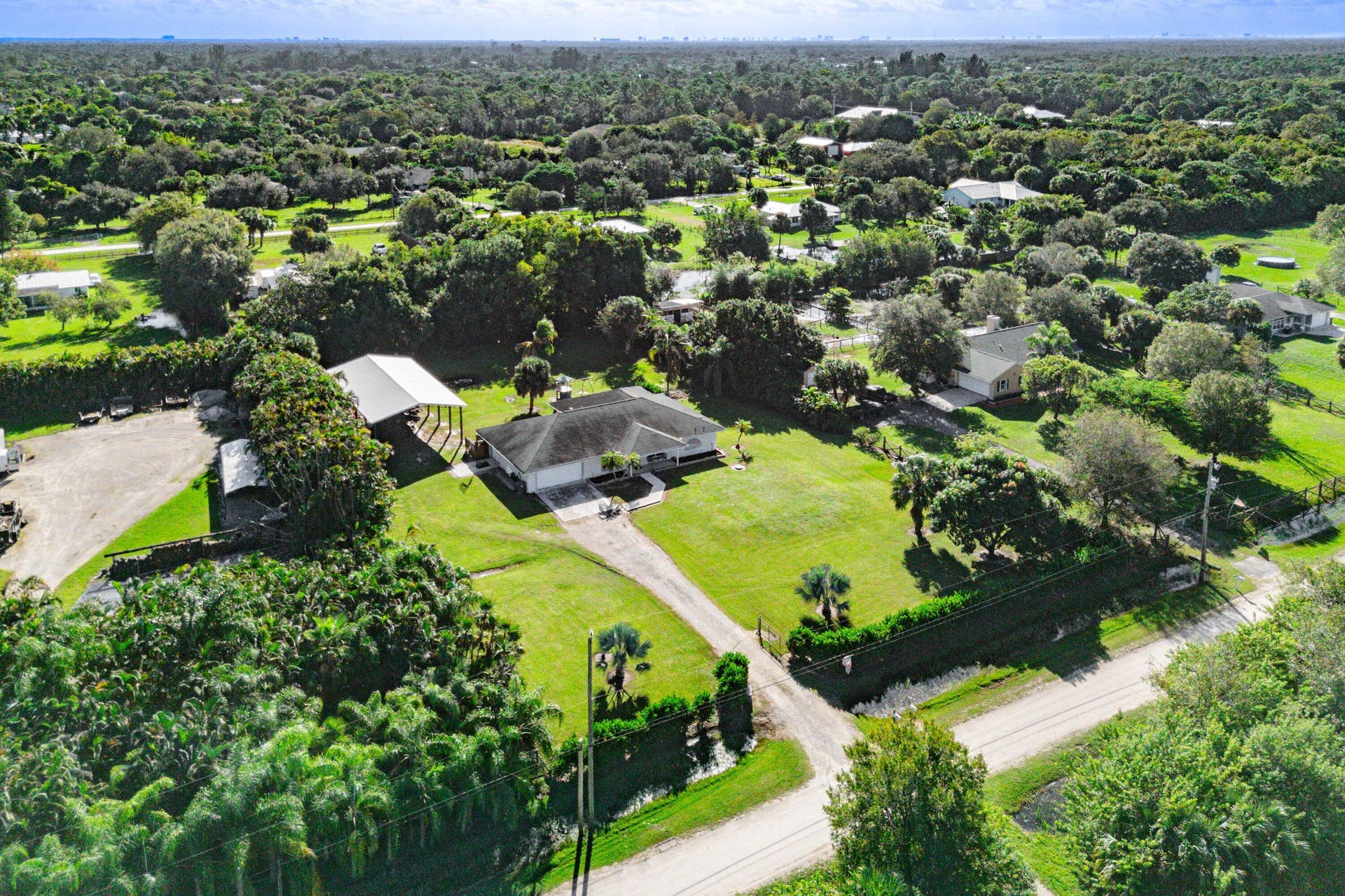 Aerial perspective of a sprawling property featuring a detached carport, expansive grounds with mature vegetation, a paved driveway, and a residential structure with a multi-hipped roofline