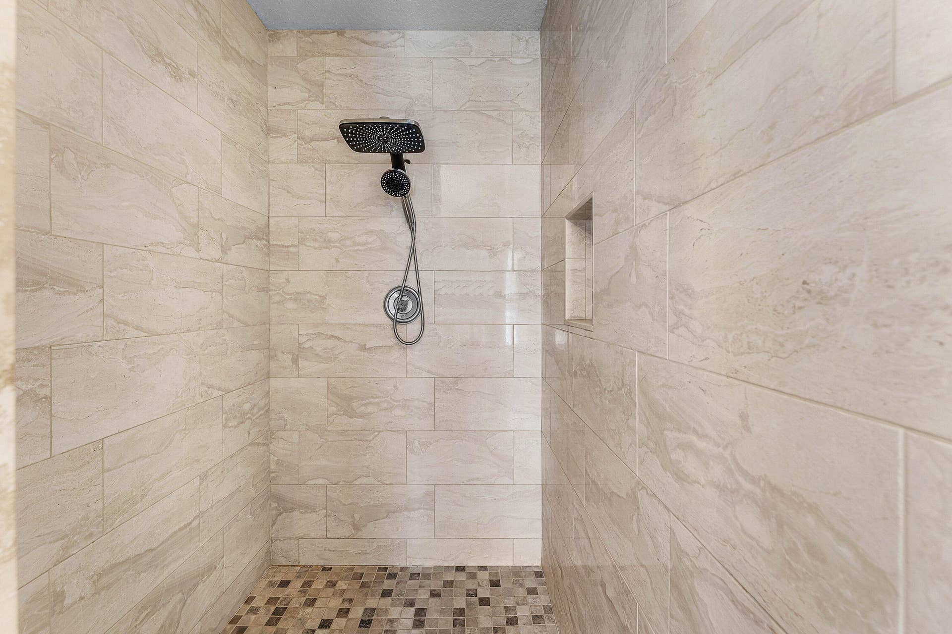Walk-in shower featuring large-format stone-finish wall tile, a square rainfall showerhead, a hand-held shower sprayer, a built-in recessed wall niche, and mosaic tile flooring