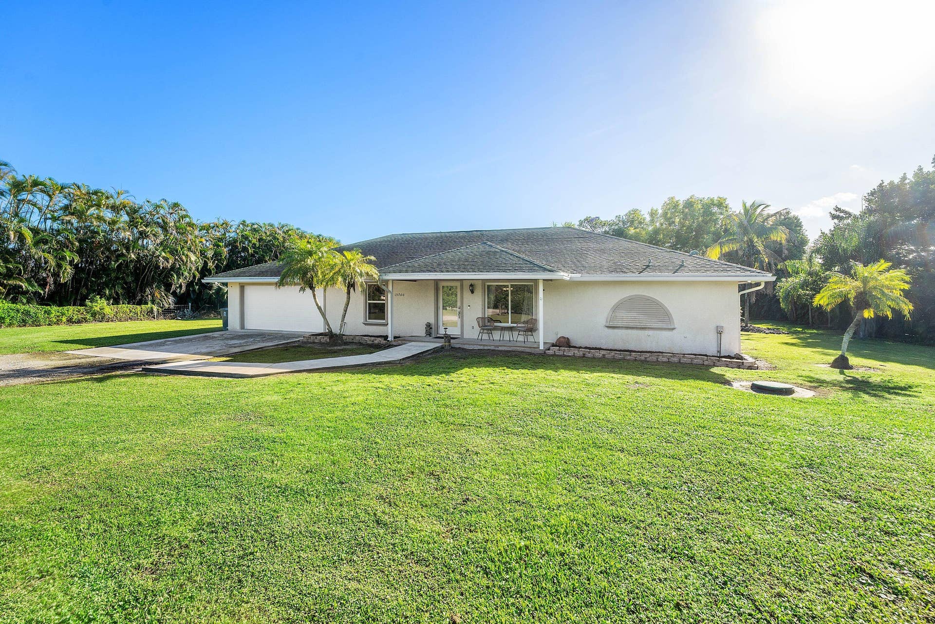 Single-story exterior featuring a light-toned stucco facade, hip roof with shingle tiles, arched window detail, concrete driveway, and sprawling lawn