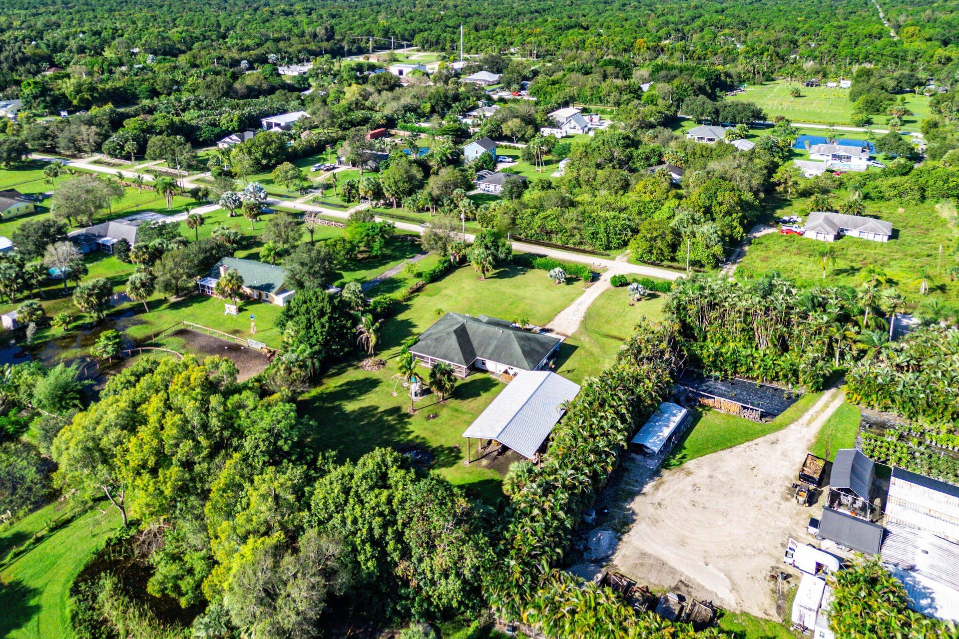 Aerial perspective of a sprawling property featuring a large residence, expansive green space, mature trees, a detached metal-roofed structure, and an unpaved driveway