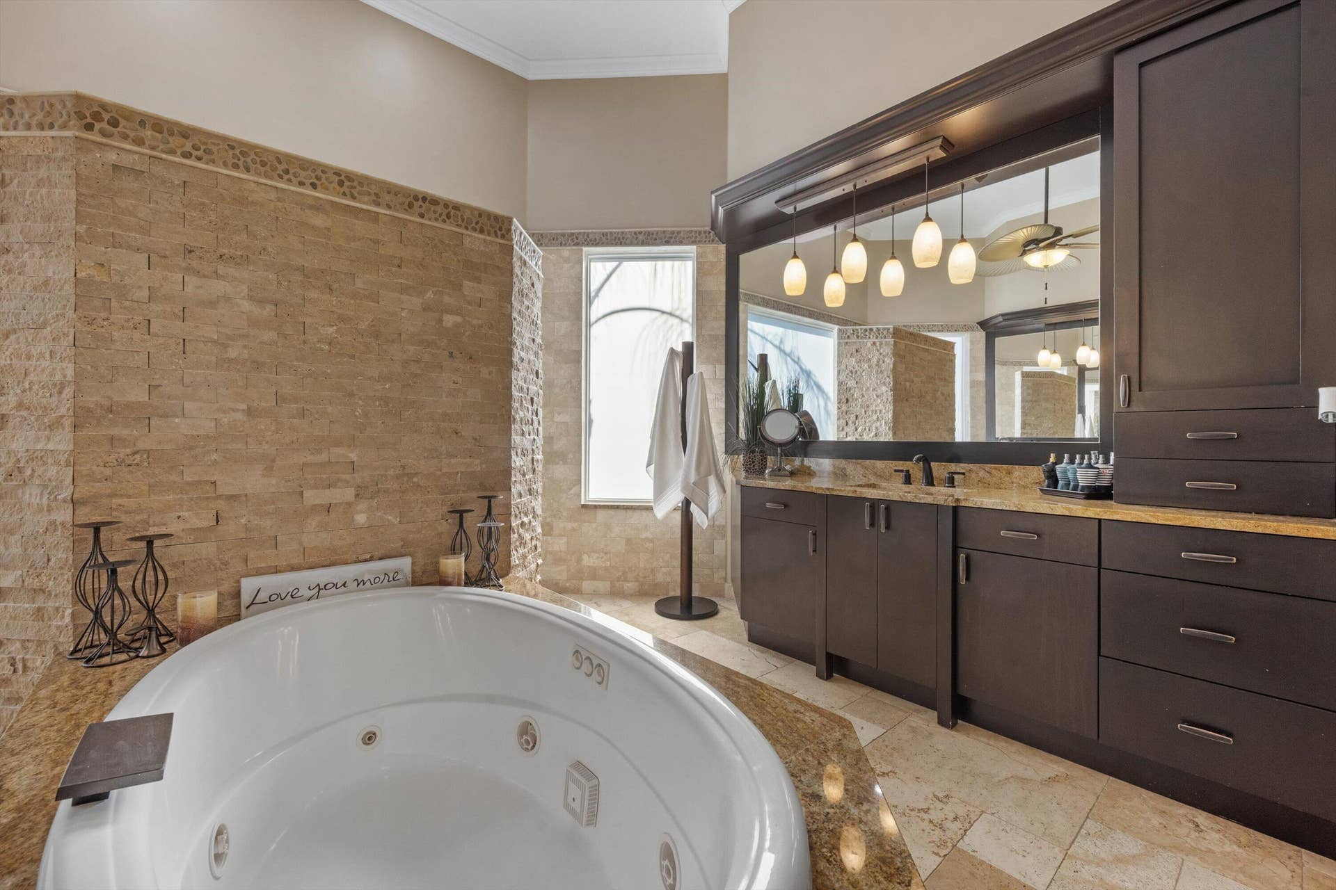 Jetted soaking tub with stone tile surround, custom dark wood cabinetry, integrated lighting, stone-finish countertop, and decorative wall stonework