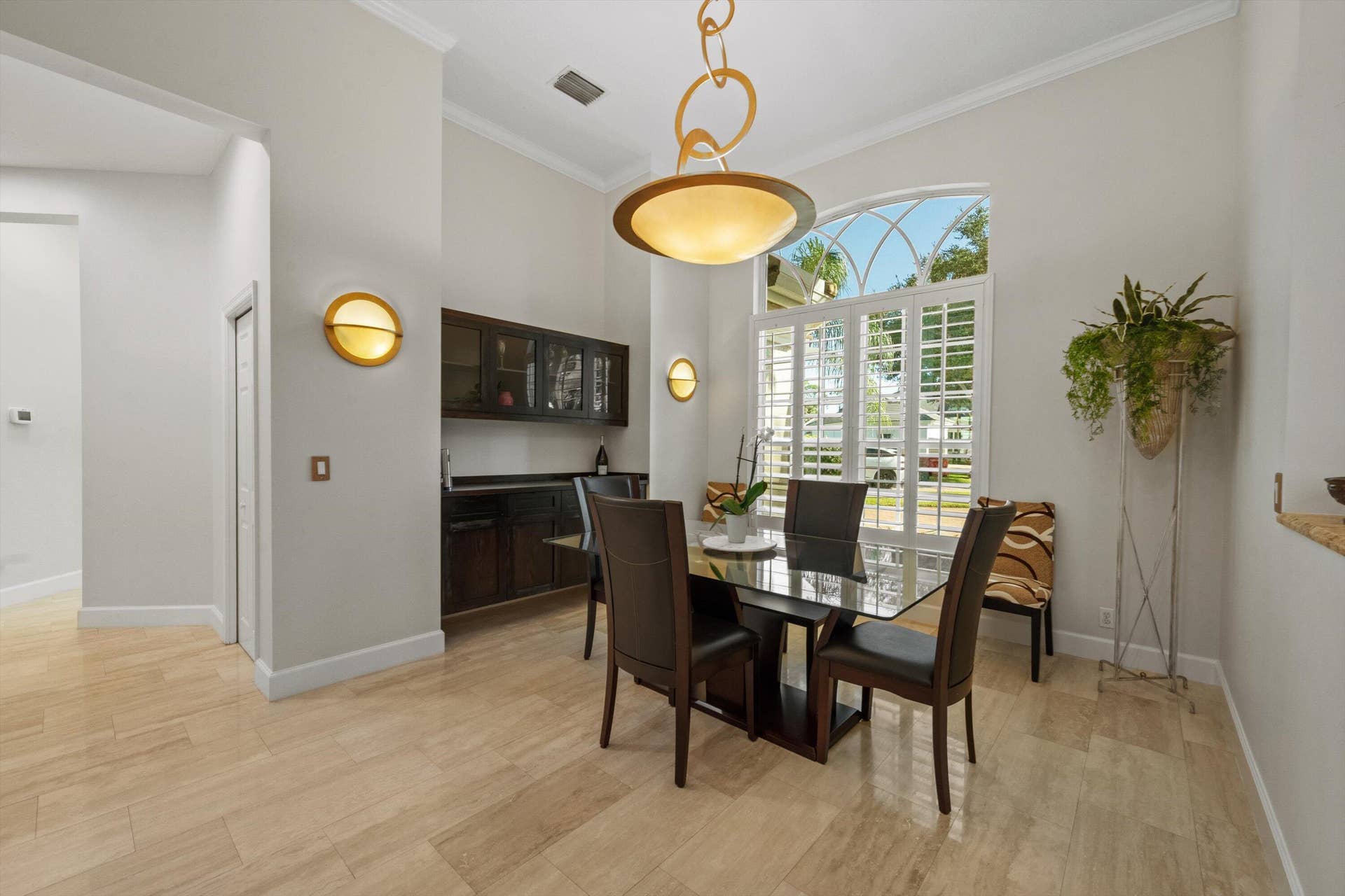 High-ceiling room featuring large arched window with plantation shutters, built-in dark wood cabinetry, polished light-tone tile flooring, and integrated wall sconces