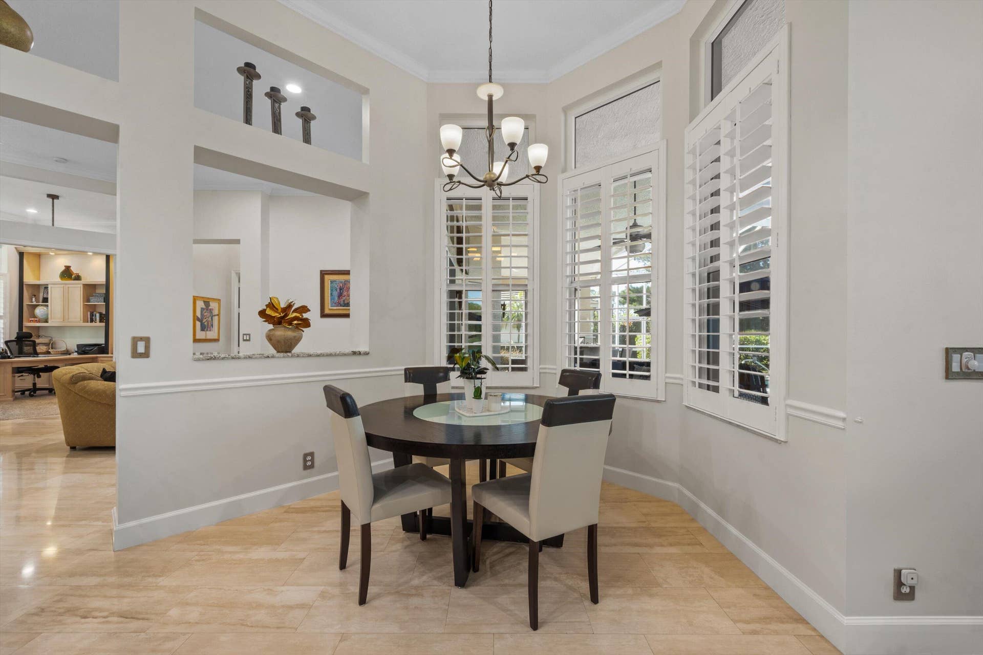 Dining area featuring light-toned tile flooring, custom plantation shutters, tiered wall niches, and a multi-arm chandelier