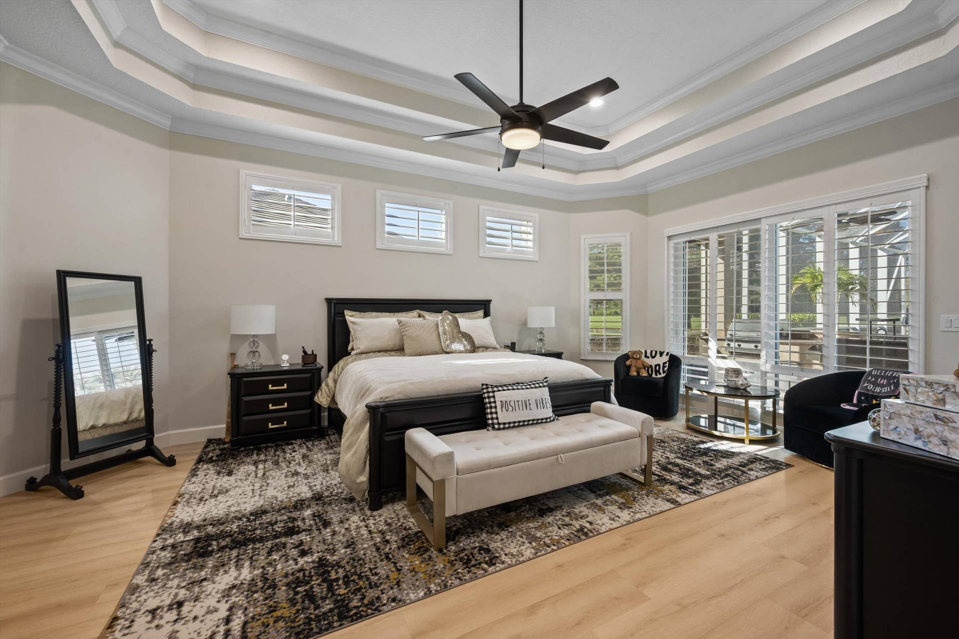 Tray ceiling with crown molding, wood-finish flooring, central ceiling fan, multiple wall-mounted plantation shutters, and sliding glass doors
