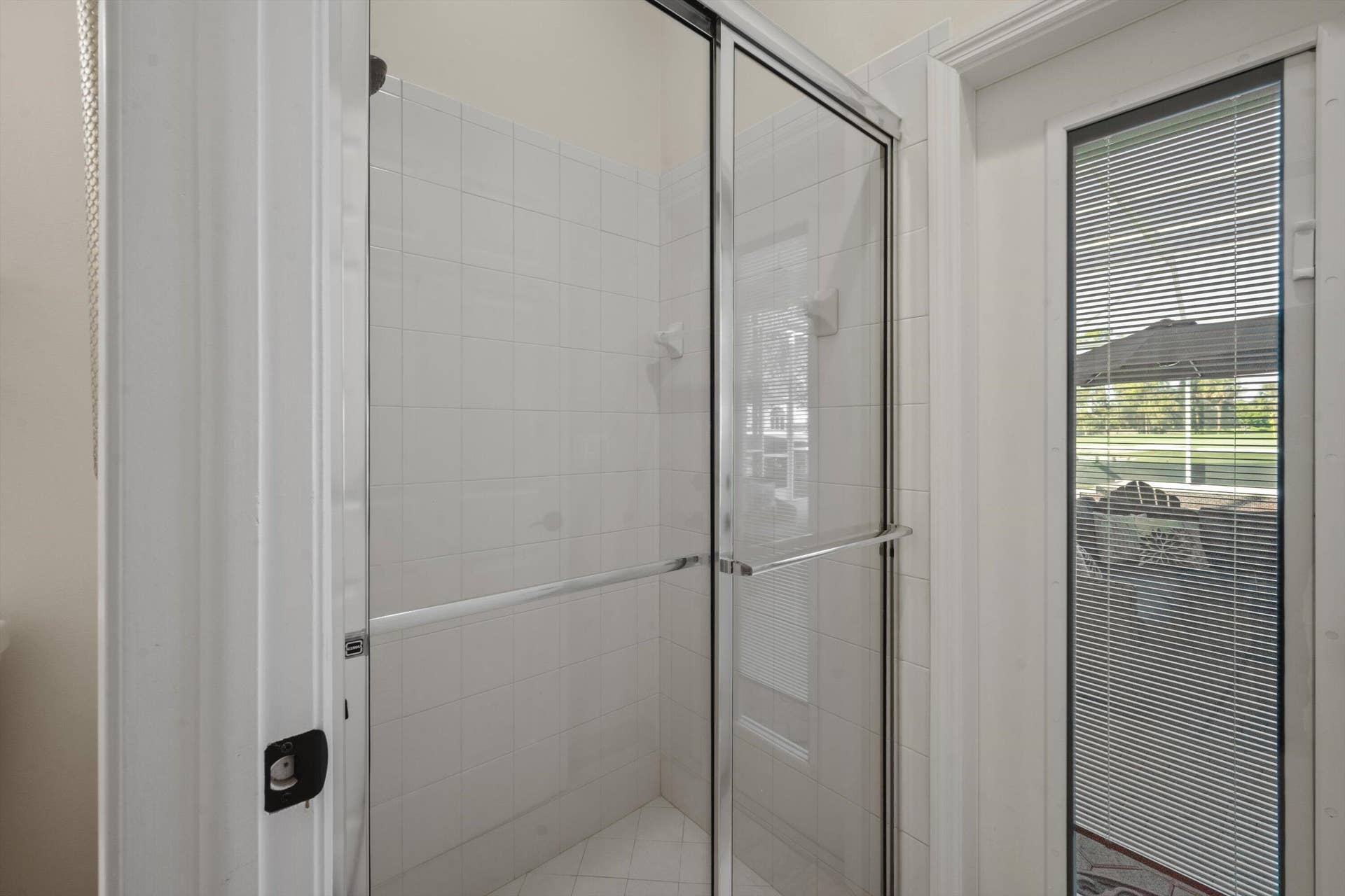 Shower enclosure featuring a chrome-framed sliding glass door, white tiled walls, and an exterior door with integrated blinds