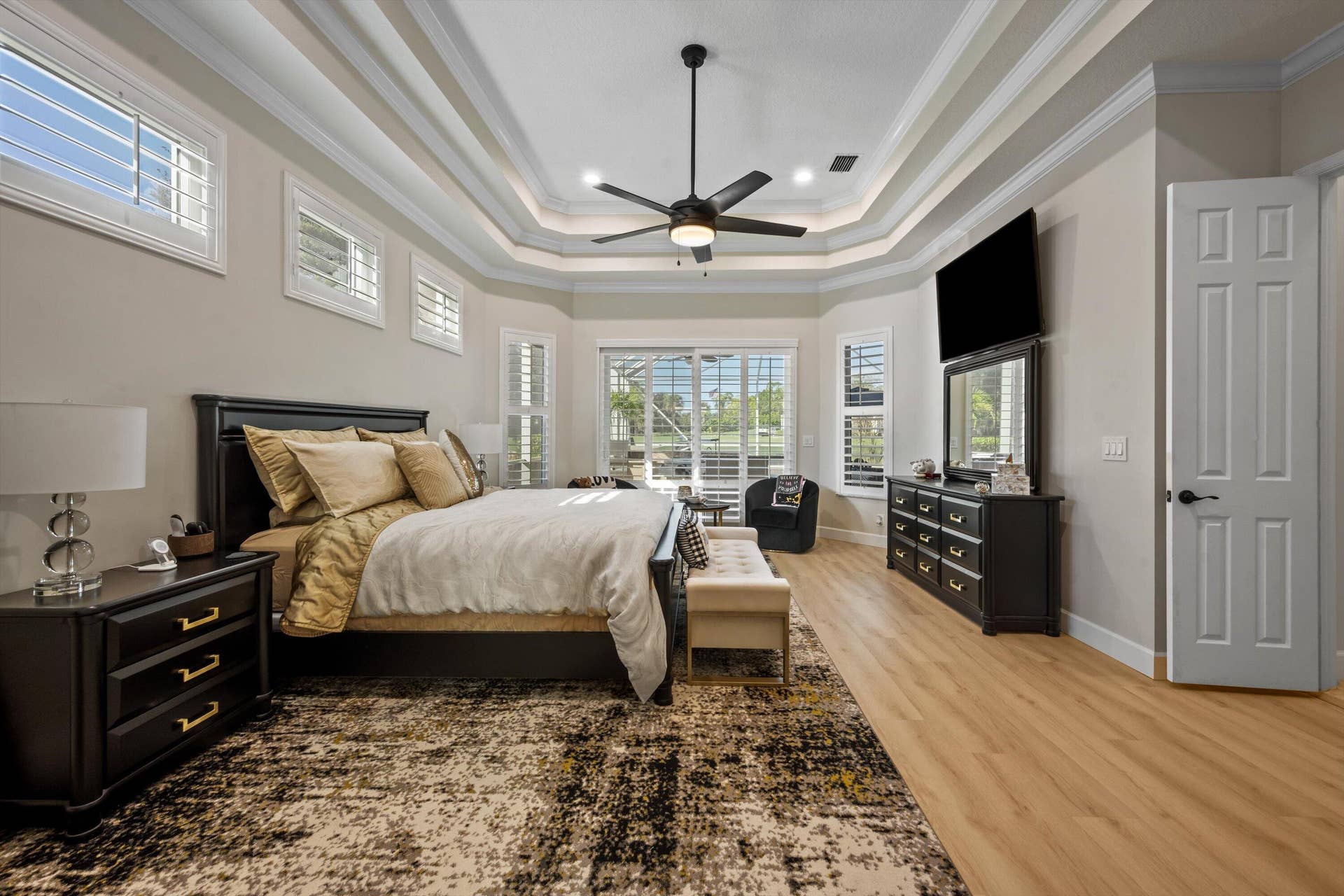 Tray ceiling with crown molding, wood-finish flooring, central ceiling fan, and multiple plantation-style shutters