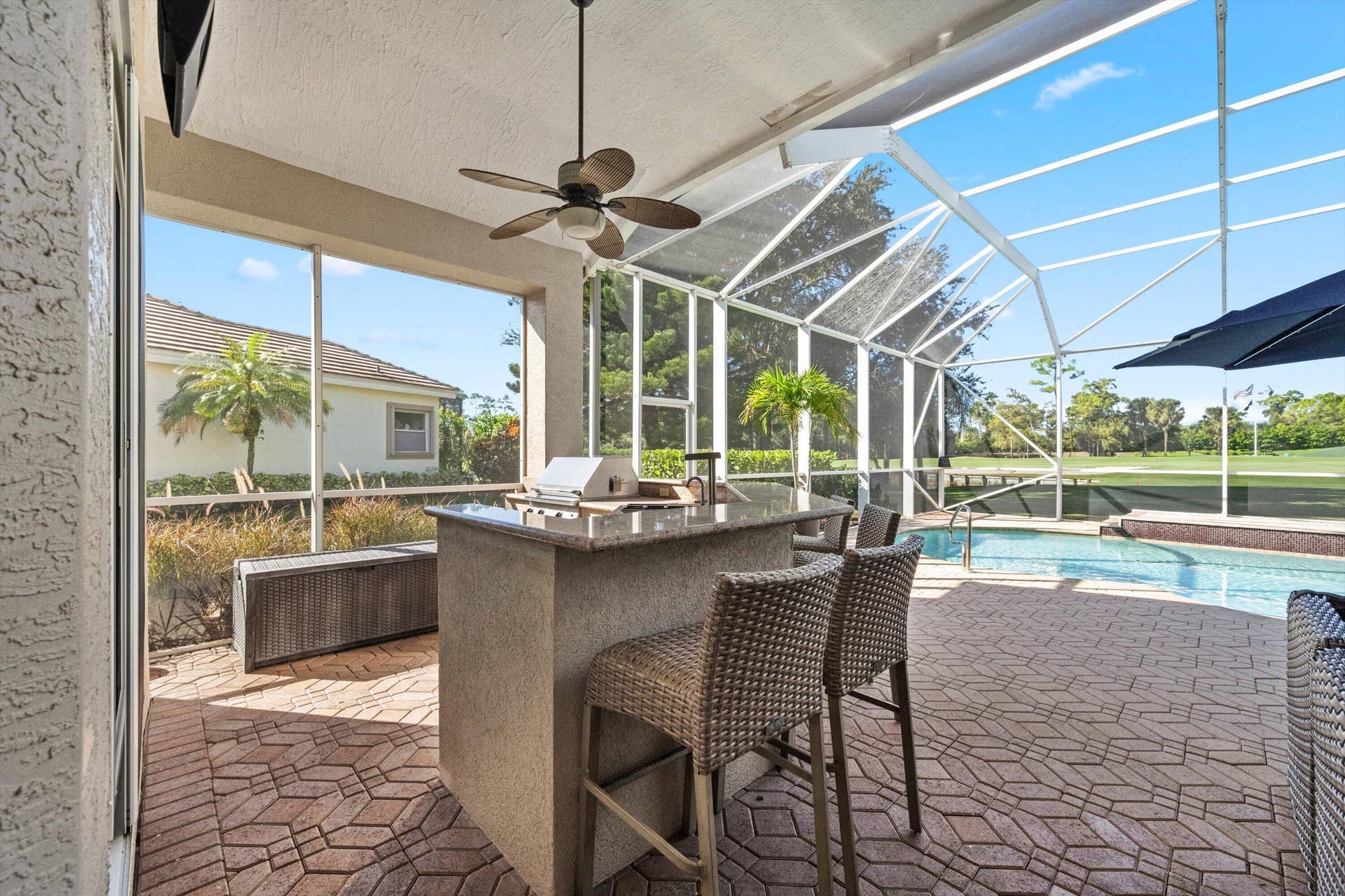 Covered lanai featuring a stone-topped outdoor kitchen, built-in grill, ceiling fan, and decorative paver flooring, fully enclosed by a screened pool cage