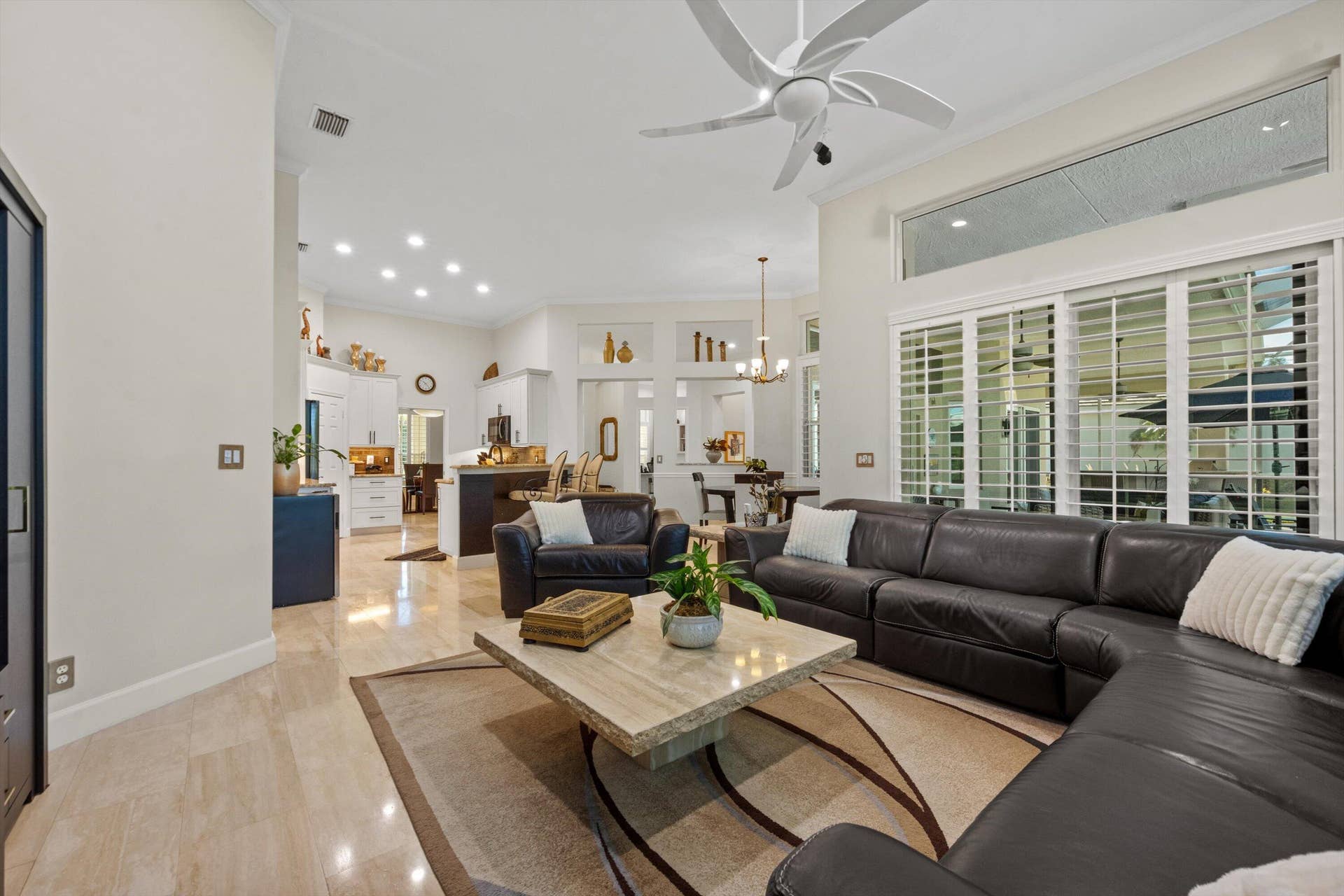 Open-concept interior featuring high ceilings, stone-finish tile flooring, recessed lighting, a multi-blade ceiling fan, and large windows with plantation shutters