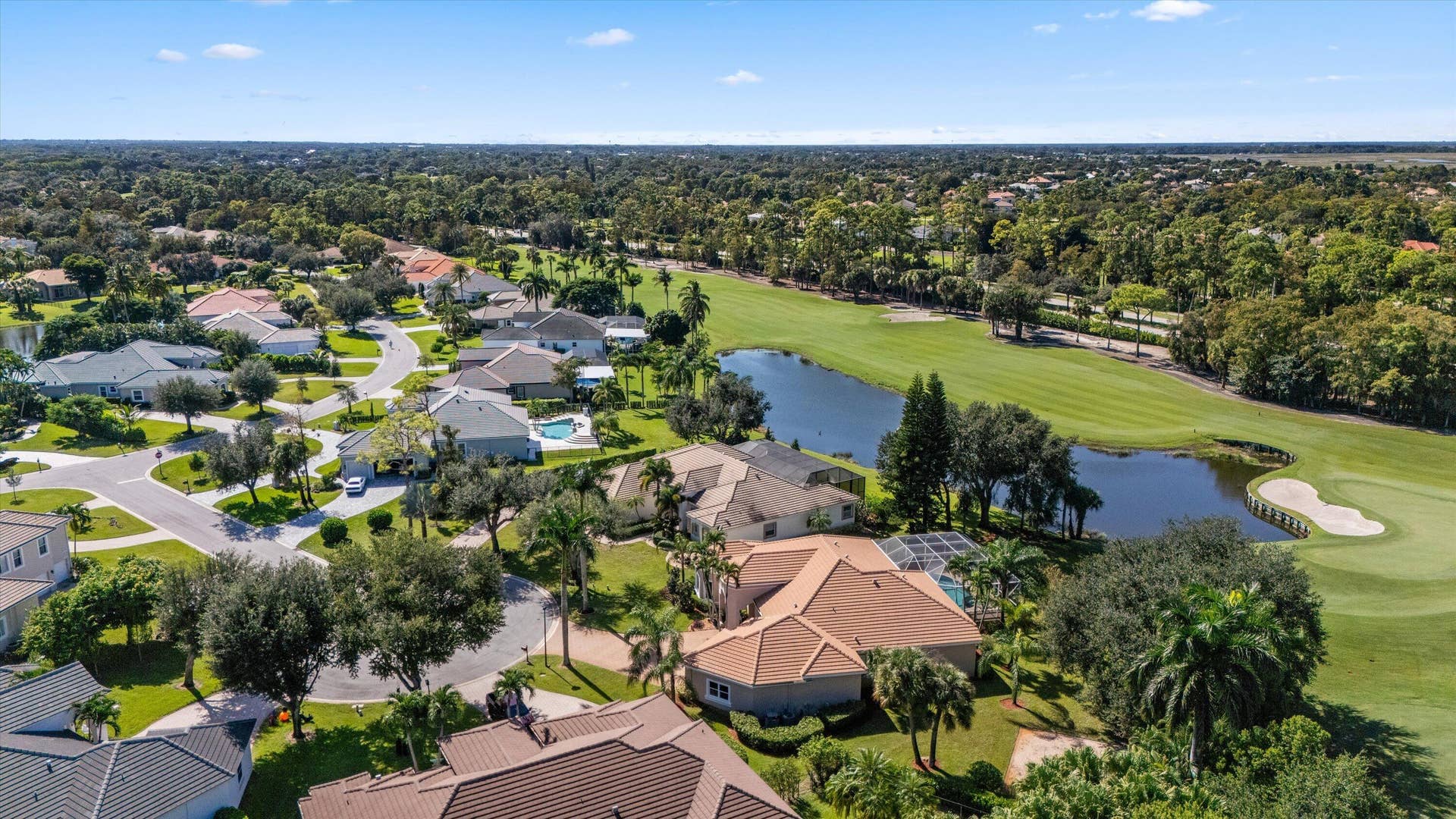 Aerial perspective featuring a multi-hipped roof residence positioned alongside a golf course fairway and a water feature, incorporating mature trees and landscape plantings within a suburban setting