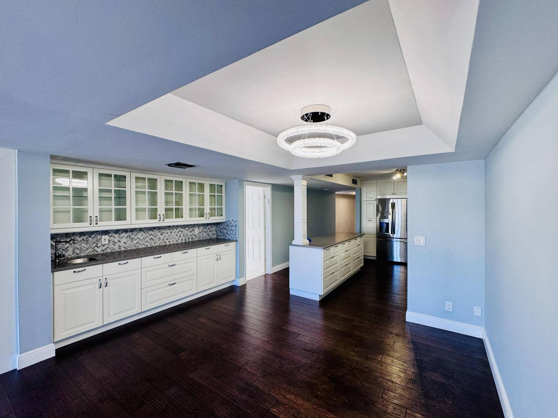 Tray ceiling with integrated circular lighting, wood-finish flooring, custom white cabinetry with glass-front uppers, herringbone-pattern tile backsplash, and dark stone-surface countertops