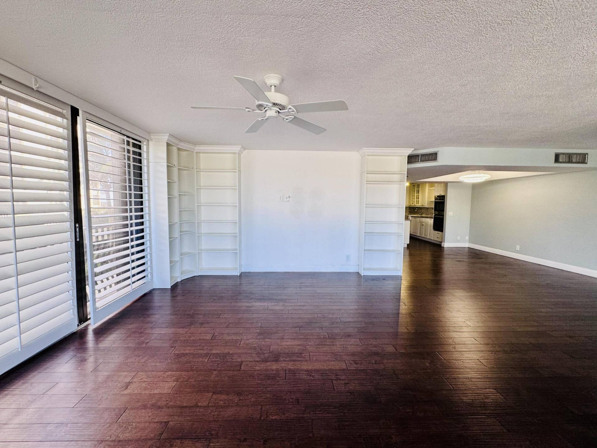 Dark-toned wood-finish flooring, white floor-to-ceiling built-in shelving, white plantation shutters, and a ceiling fan