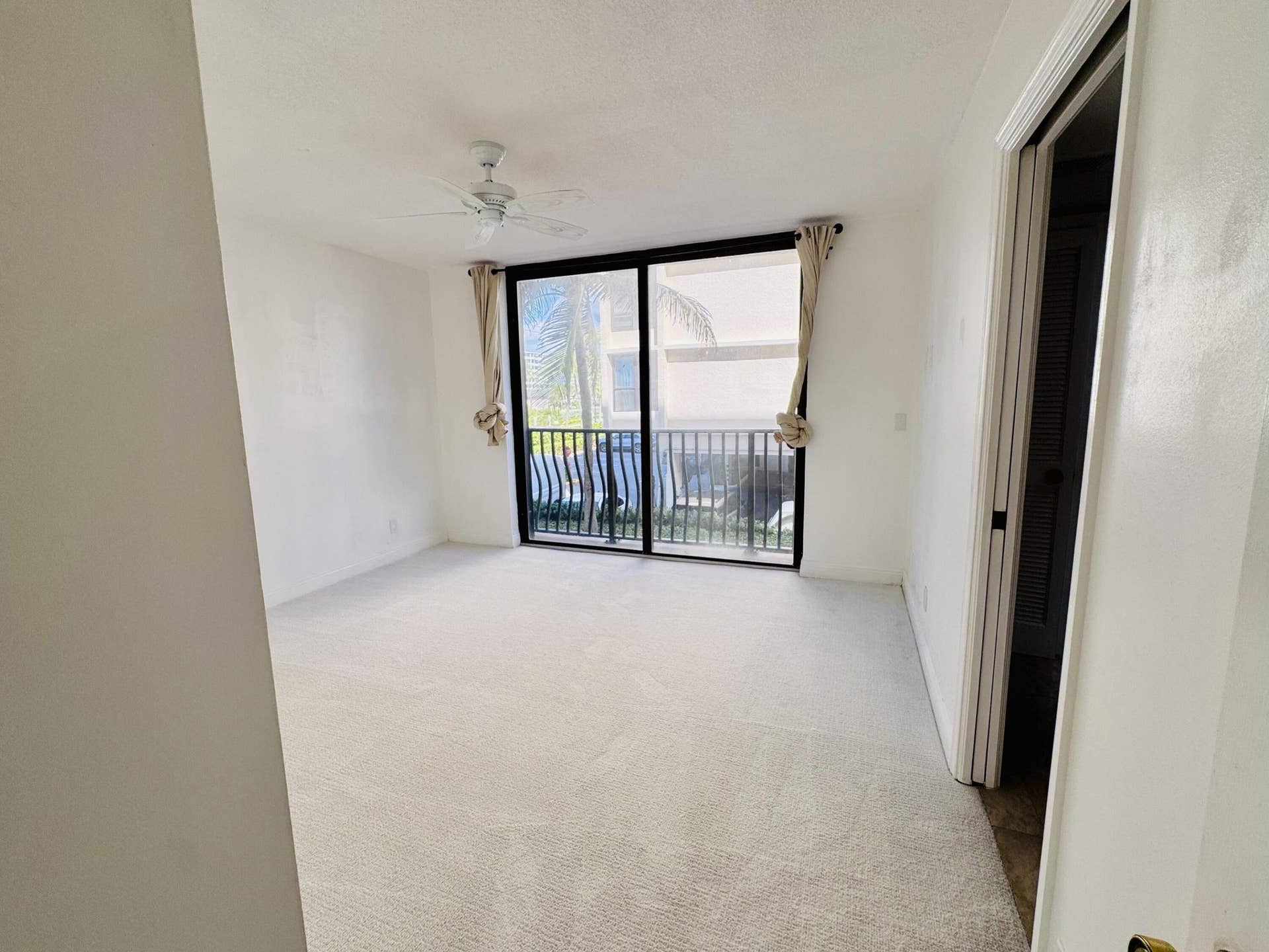 Neutral-toned wall-to-wall carpeting, ceiling-mounted paddle fan, and large floor-to-ceiling glass sliding doors opening to a private balcony with metal railing