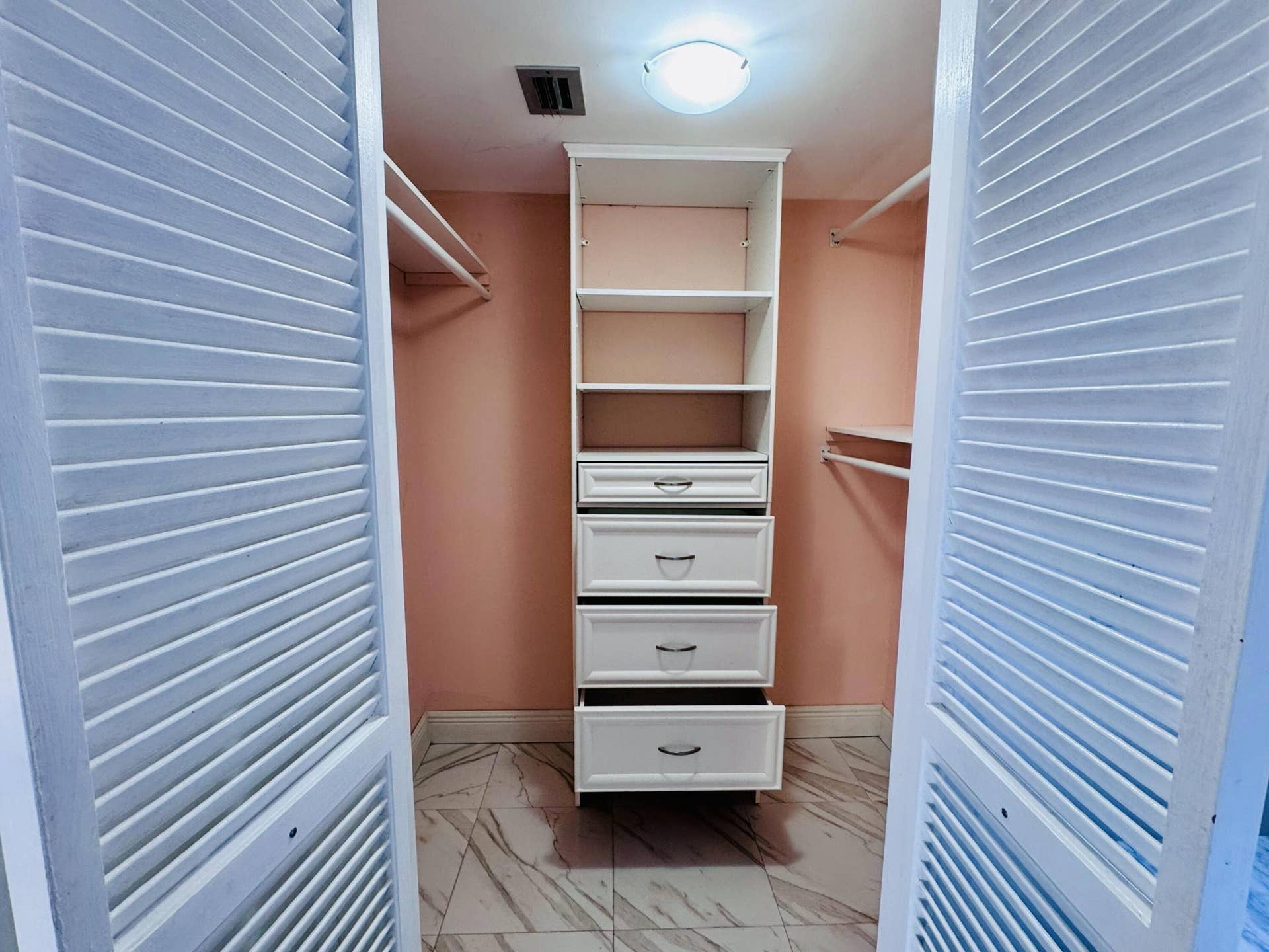Walk-in closet featuring white louvered bifold doors, built-in shelving, a four-drawer storage unit with brushed metal hardware, dual hanging rods, and patterned tile flooring