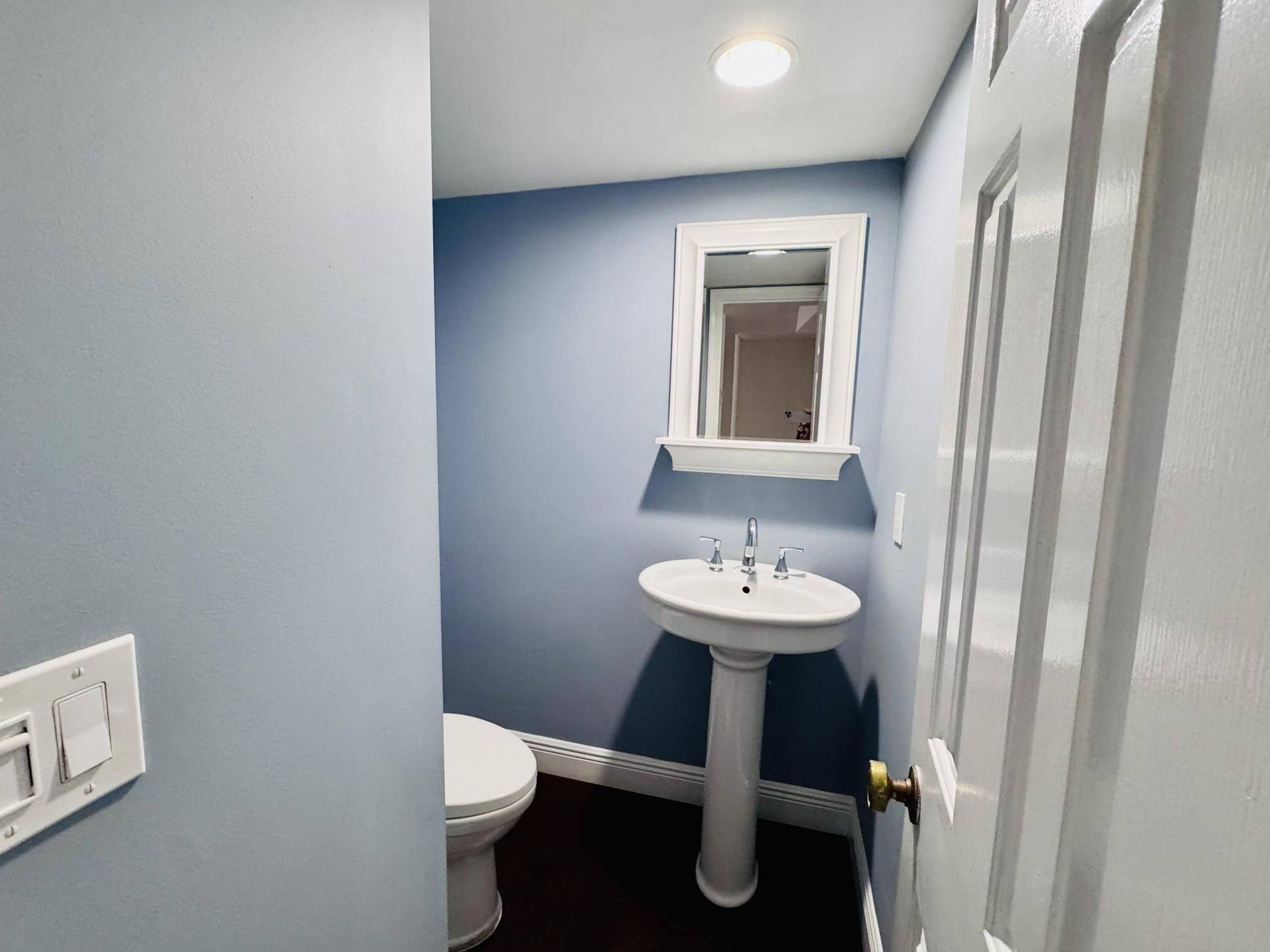 White porcelain pedestal sink with a dual-handle chrome faucet, framed wall mirror, and dark wood-finish flooring