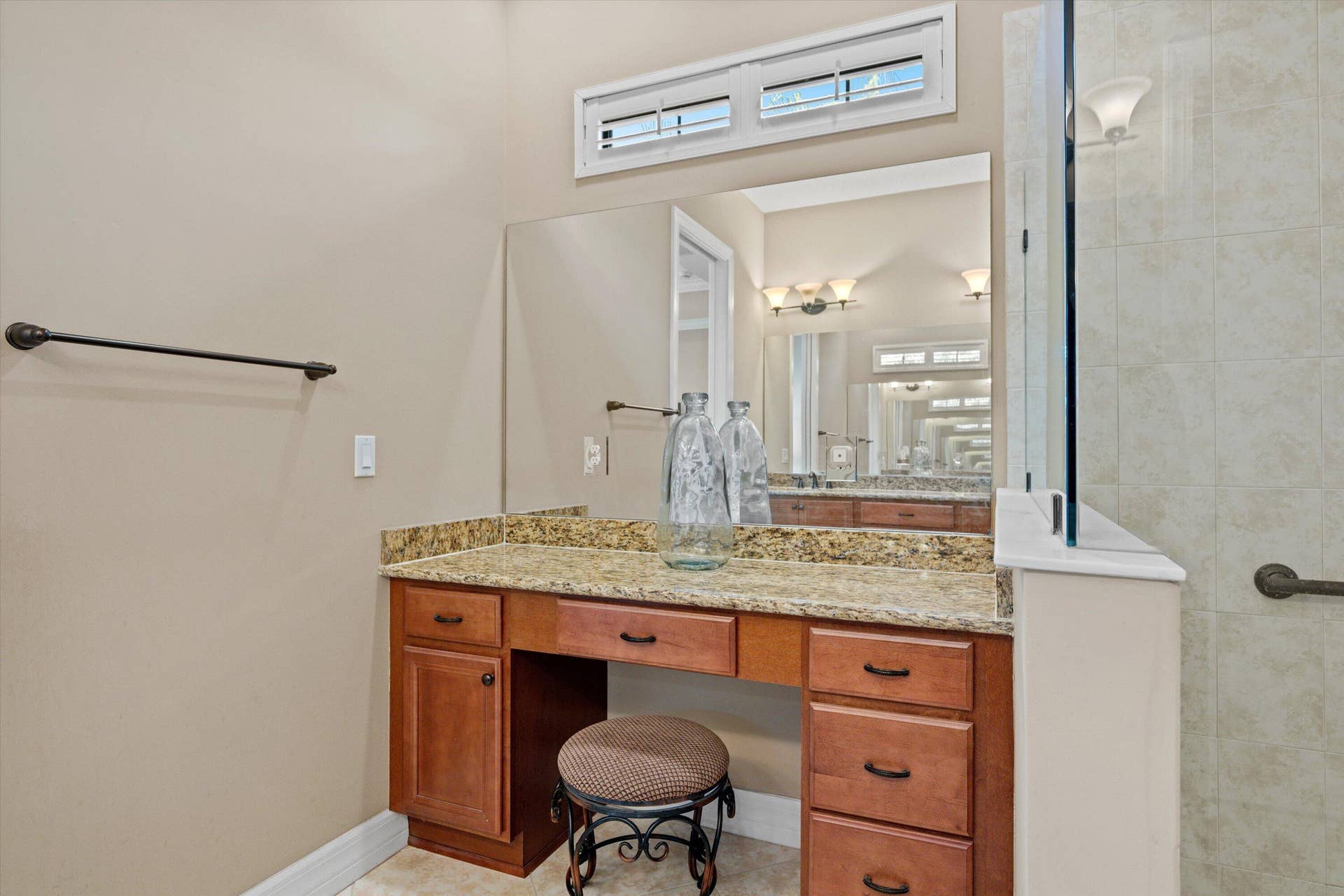 Built-in vanity featuring stone-finish countertop, wood cabinetry, large vanity mirror, transom window, and tile-surround shower