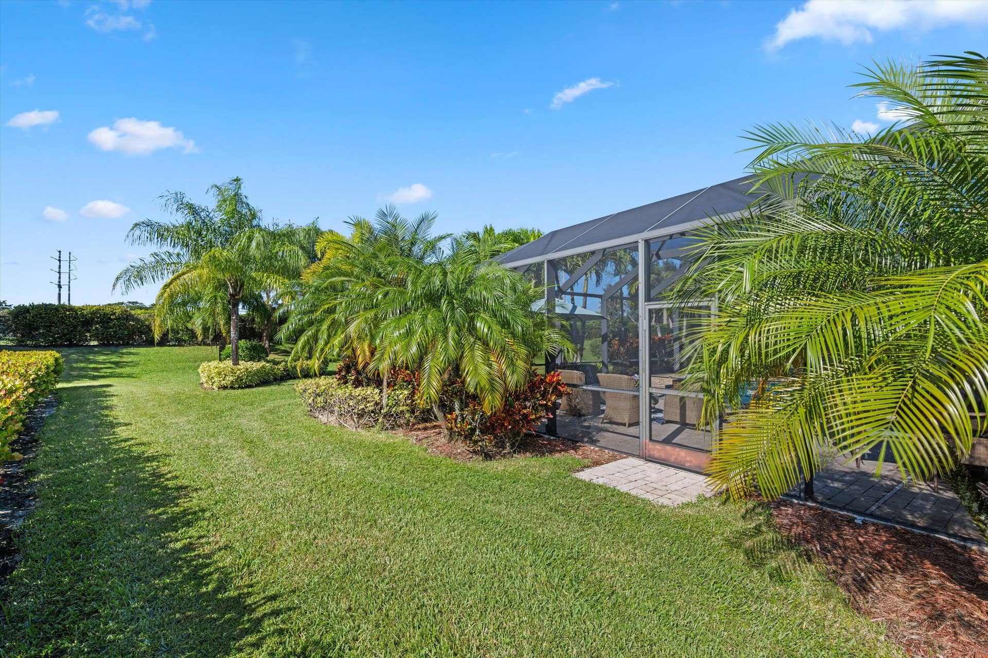 Screened-in patio enclosure featuring paver accent landing, surrounded by mature palms, manicured lawn, and professional landscape shrubbery