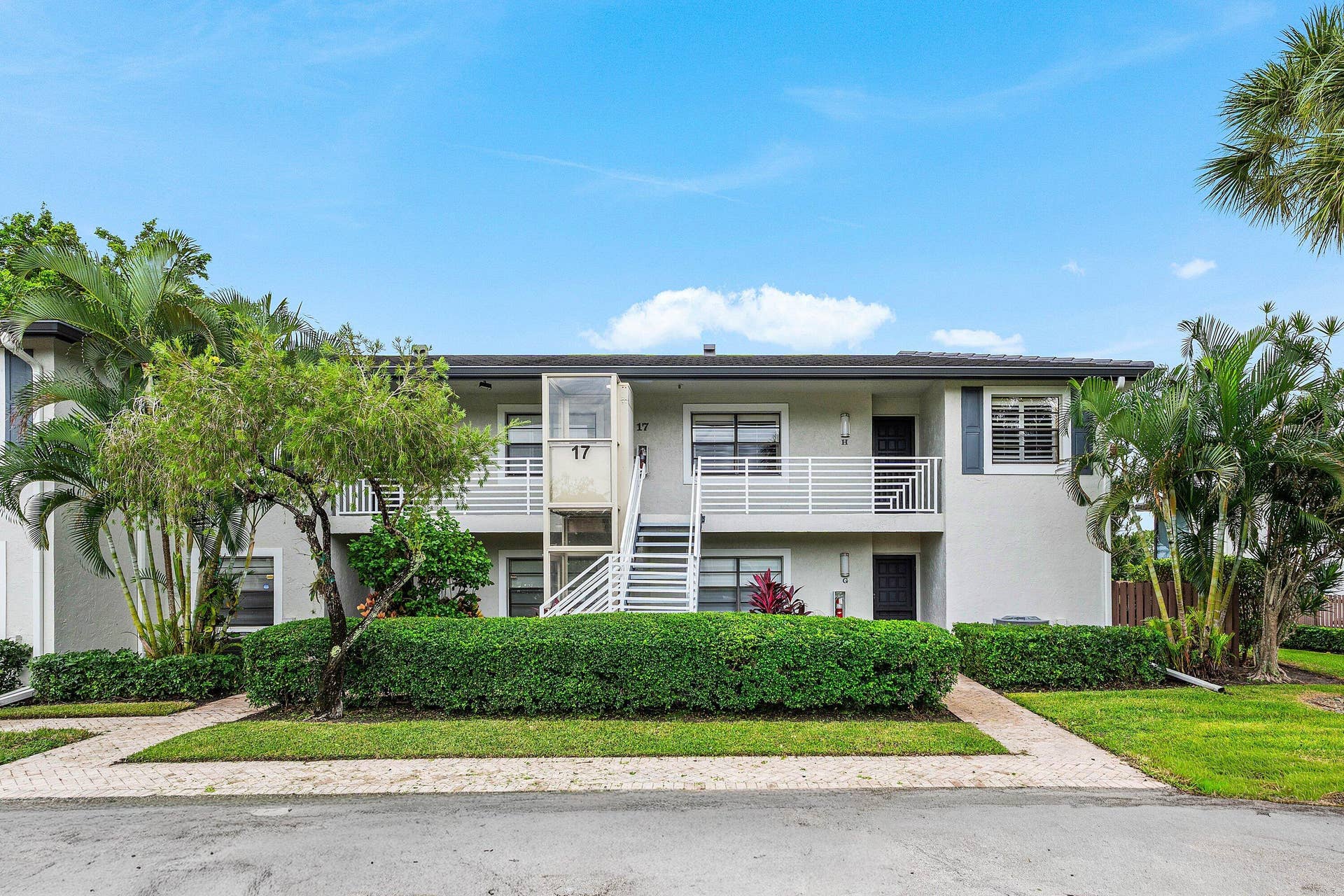 Two-story exterior featuring a light-toned stucco faade, white horizontal-slat balcony railings, metal exterior staircase, and paved walkway