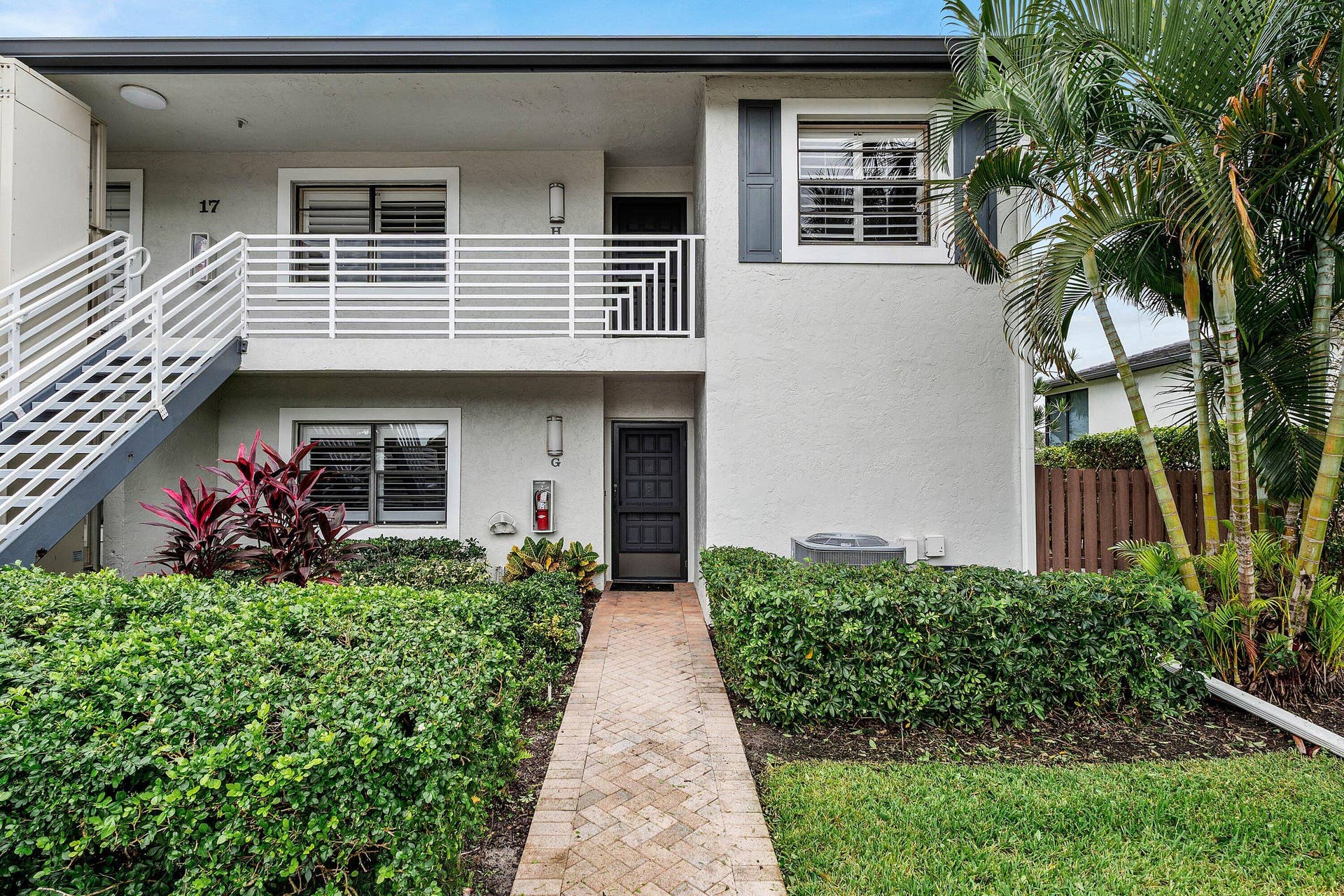 Two-story building exterior featuring a textured stucco facade, a brick-paved walkway, a second-level balcony with white horizontal railings, and mature landscaping