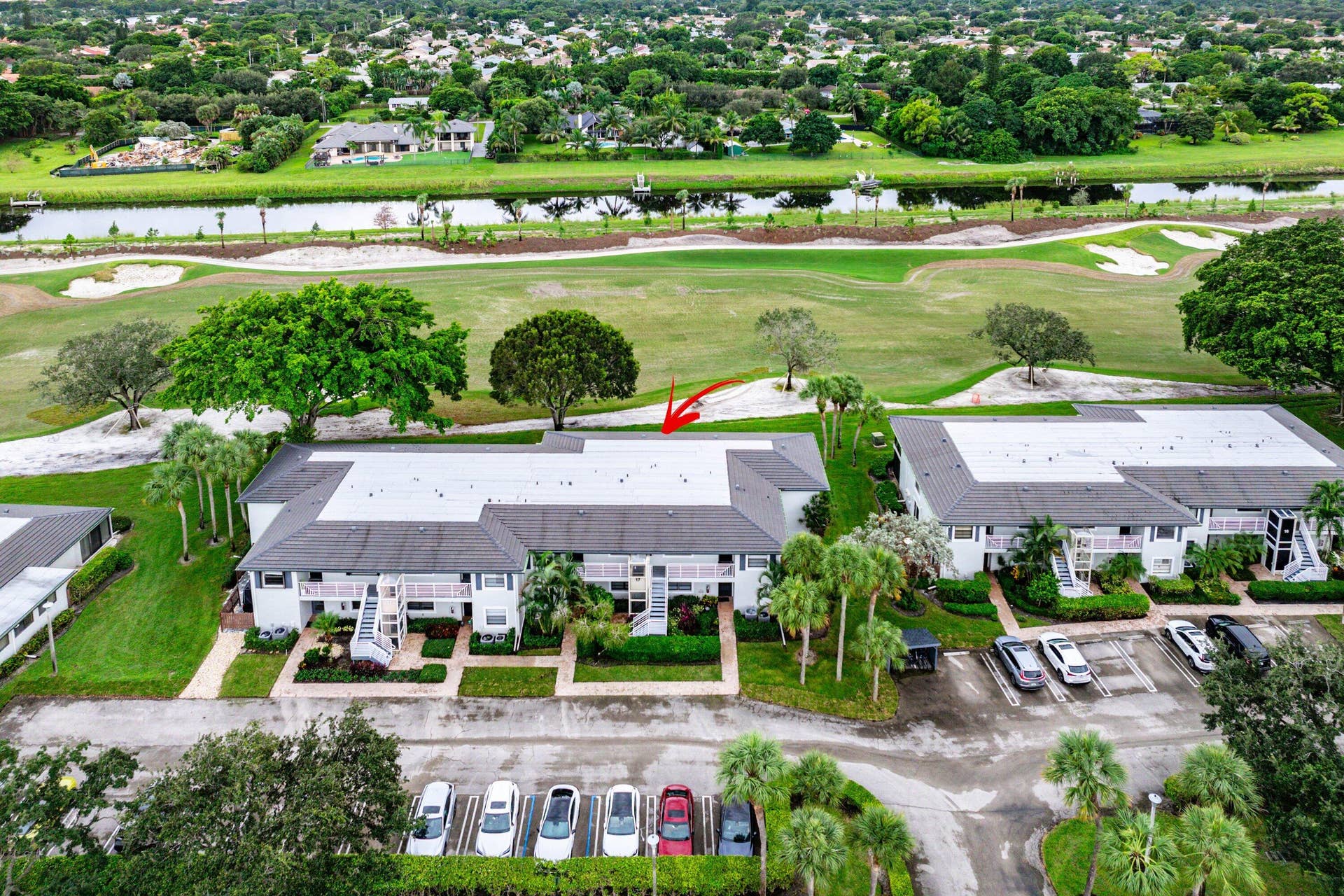 Aerial perspective of a multi-unit residential building featuring a multi-hipped roof, paved parking area, landscaping, and immediate proximity to a golf course and canal