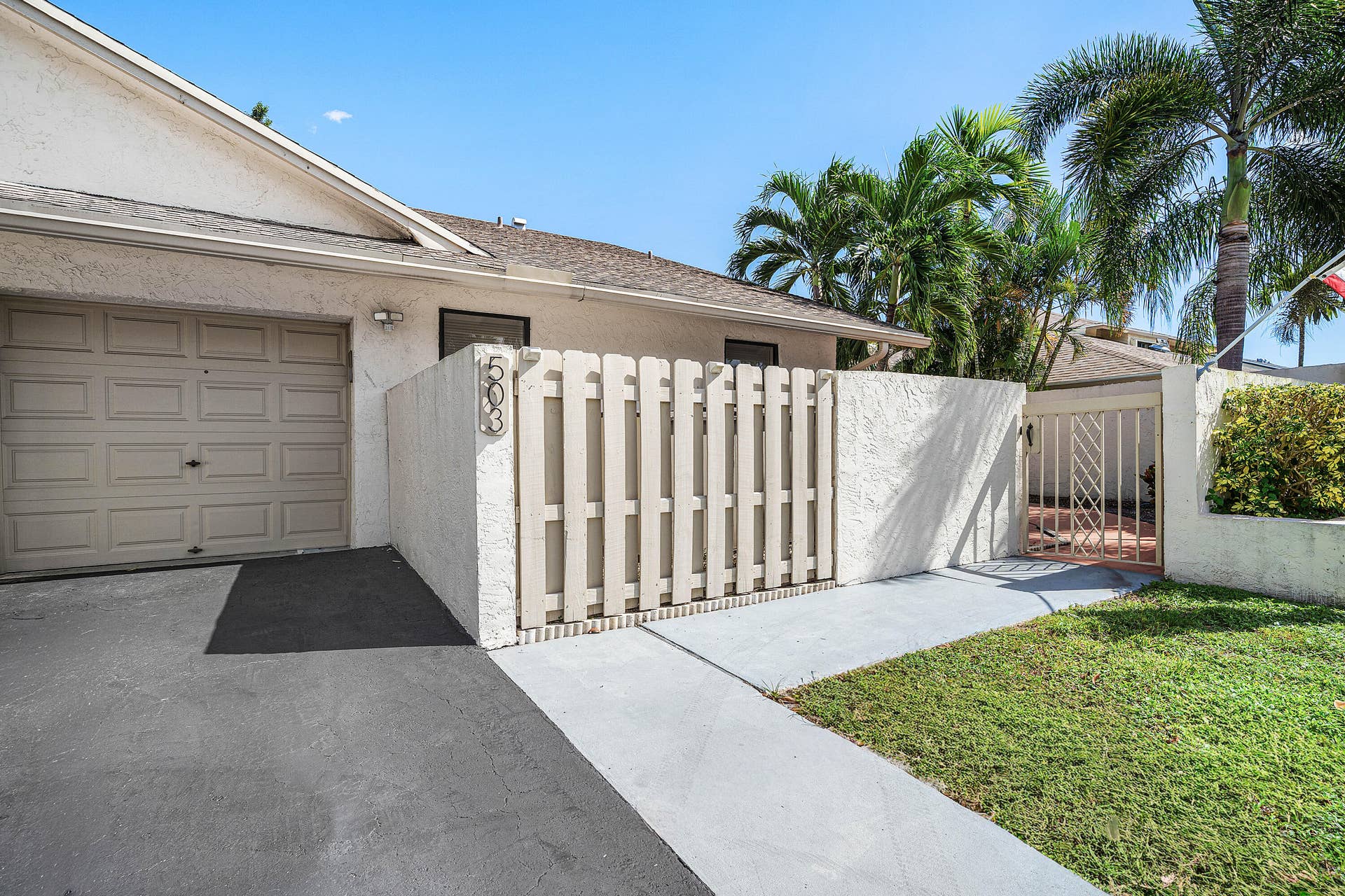 503 SW 29TH Avenue, Delray Beach, FL 33445 Stucco exterior featuring an attached one-car garage, private enclosed courtyard with vertical slat fencing, paved walkway, and manicured lawn