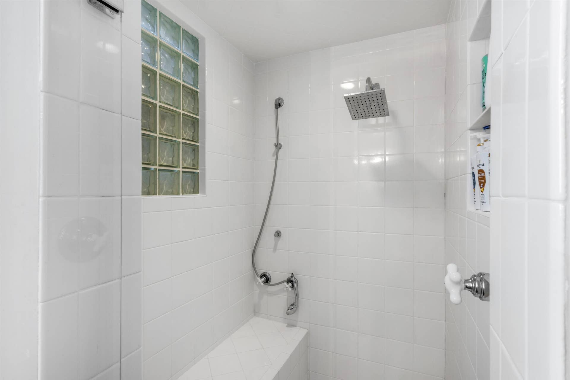419 E Atlantic Avenue, Delray Beach, FL 33483 Walk-in shower featuring white tile surround, glass block accent window, rainfall showerhead with handheld attachment, built-in bench seating, and recessed storage niche