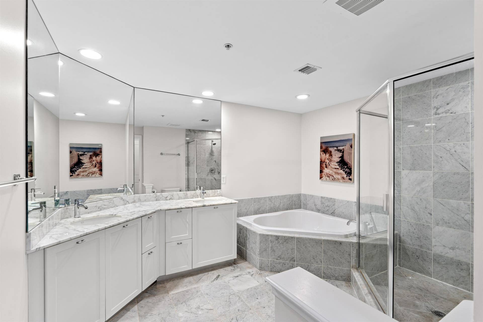 White cabinetry with stone countertops, expansive wall-to-wall mirror, corner soaking tub, stone-tiled walk-in shower with glass enclosure, and stone flooring