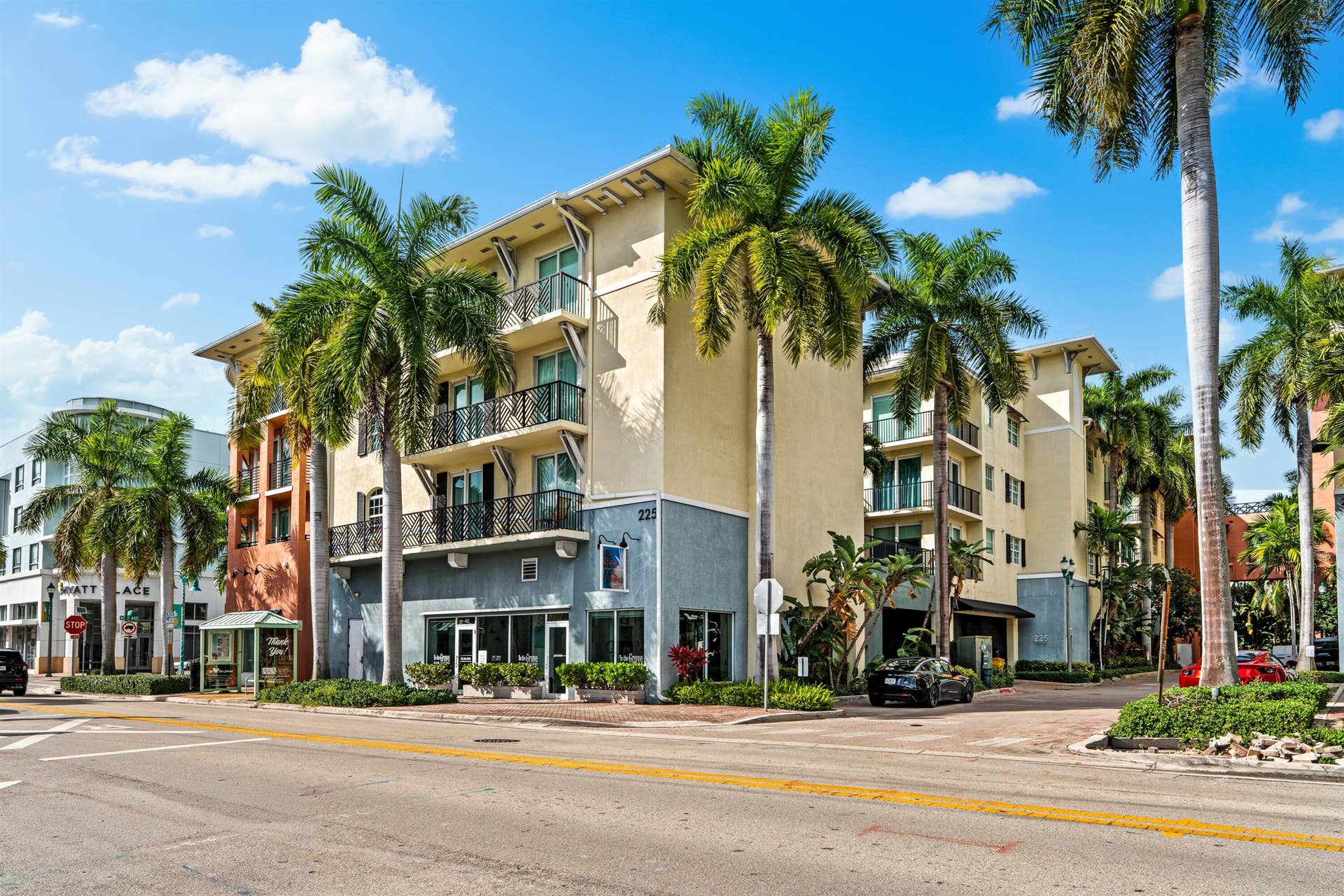 Multi-story stucco building exterior featuring metal balcony railings, prominent palm tree landscaping, and floor-to-ceiling ground-level windows