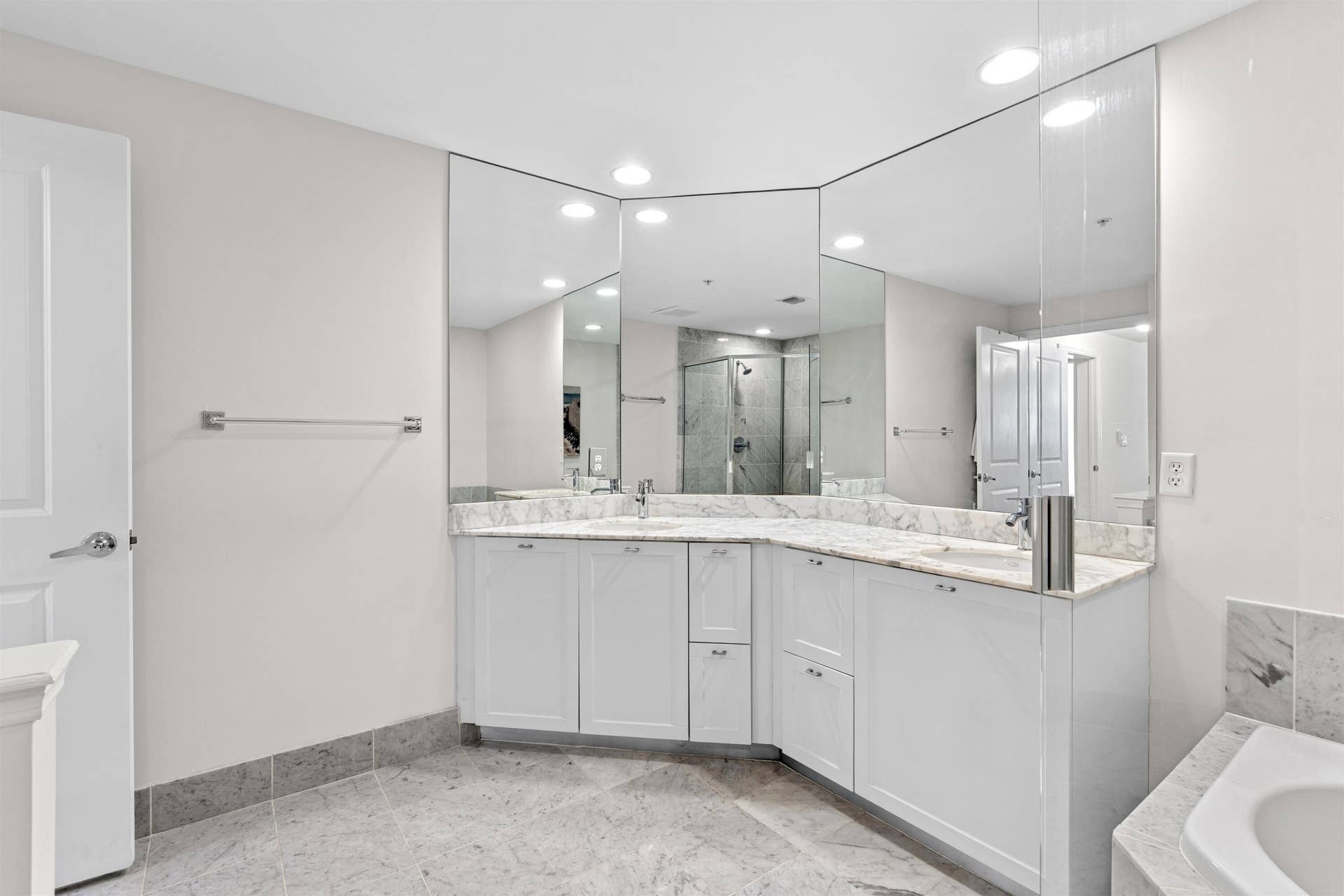 Corner vanity with white cabinetry, stone-surface countertop, dual integrated sinks, and expansive multi-panel wall mirror, featuring tile flooring and recessed lighting