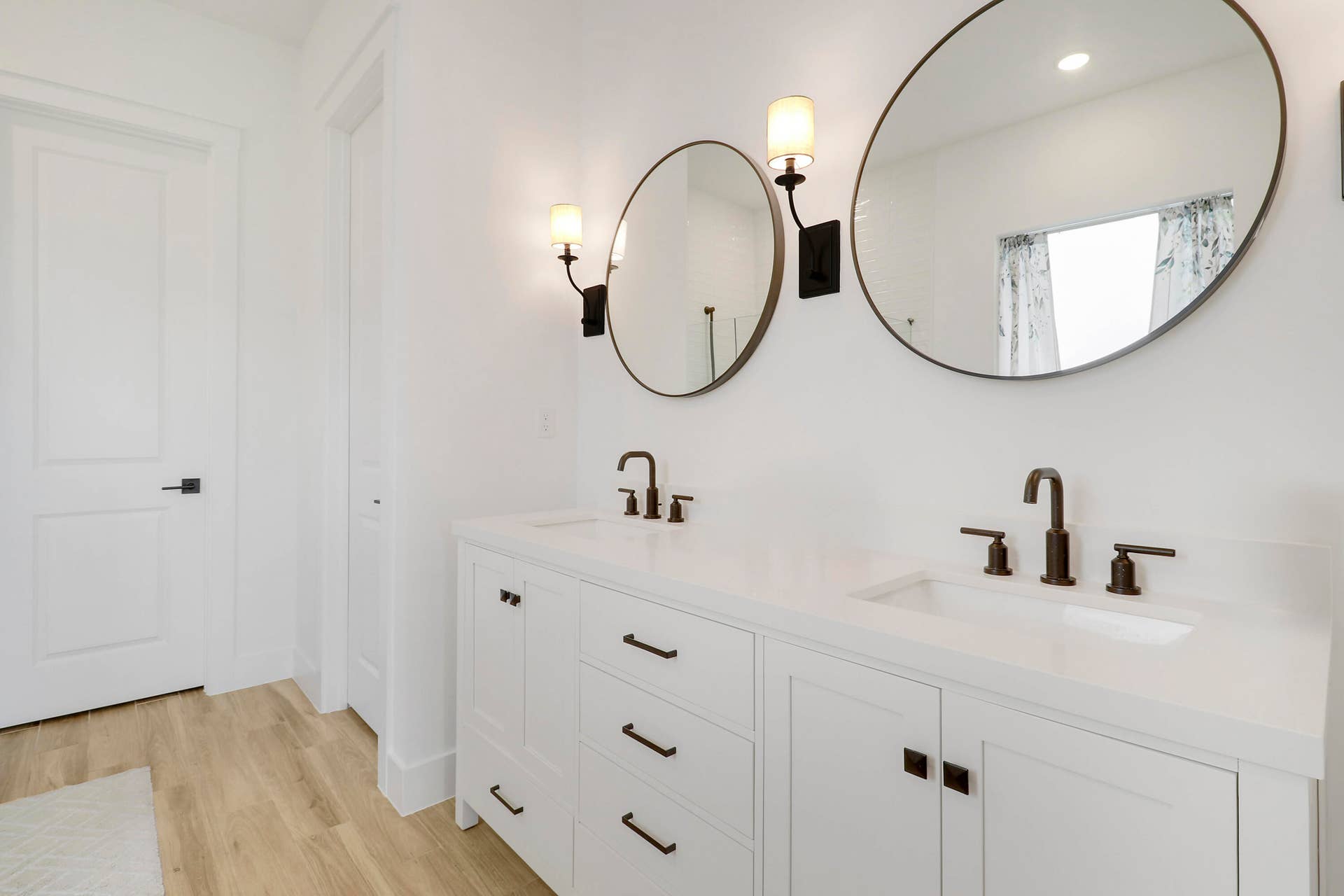 Dual-sink vanity featuring a white composite countertop, undermount sinks, matte black widespread faucets, and shaker-style cabinetry with matte black hardware, complemented by round-frame mirrors and wall-mounted sconces