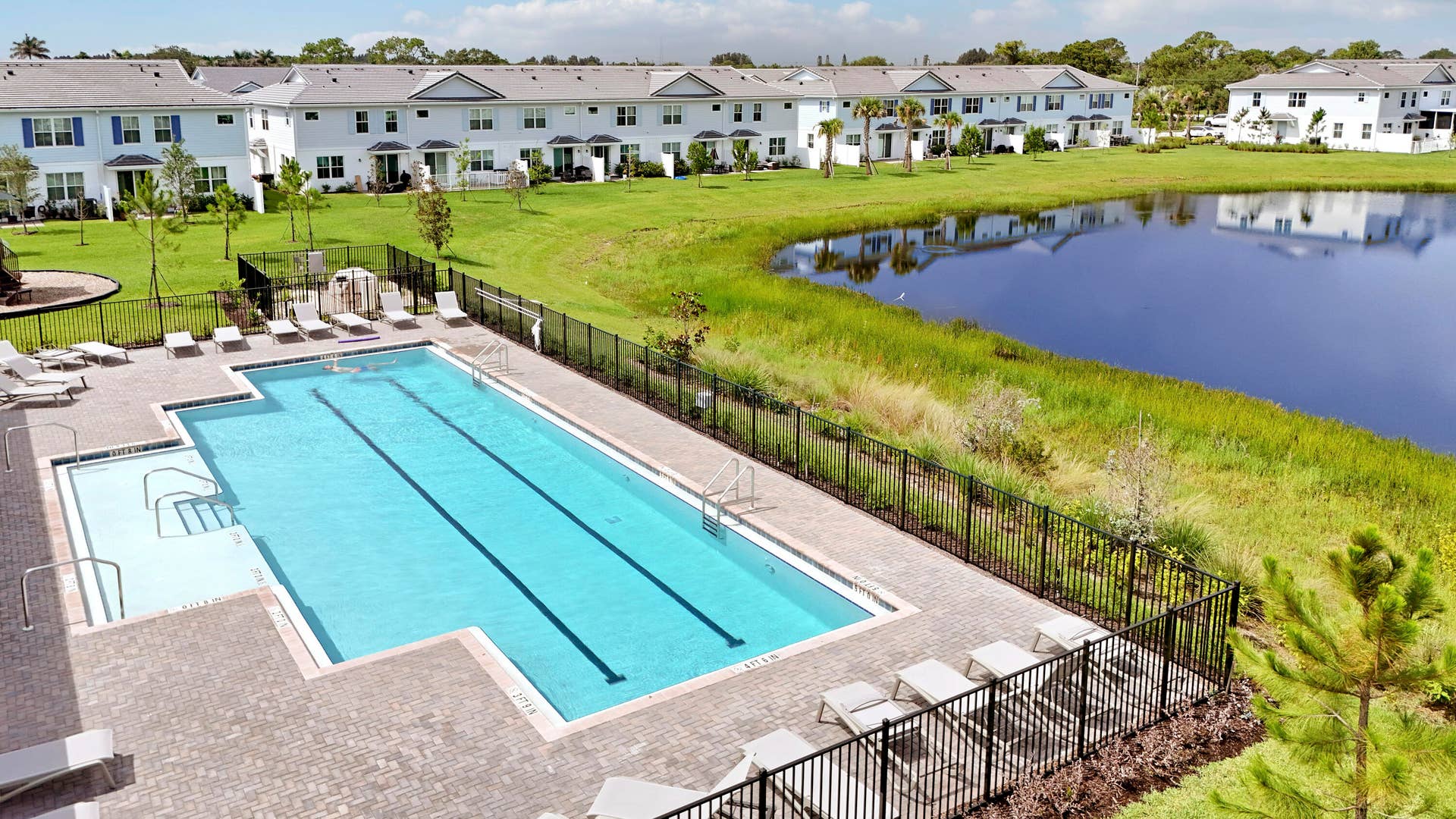 Community swimming pool with lap lanes and shallow entry, surrounded by paver decking and enclosed by black metal fencing, situated near a retention pond.