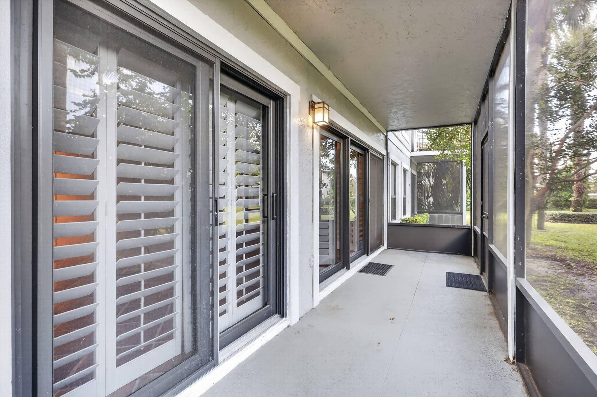 Screened-in porch featuring concrete flooring, dark metal framing, sliding glass doors with white plantation shutters, and a wall-mounted exterior light fixture