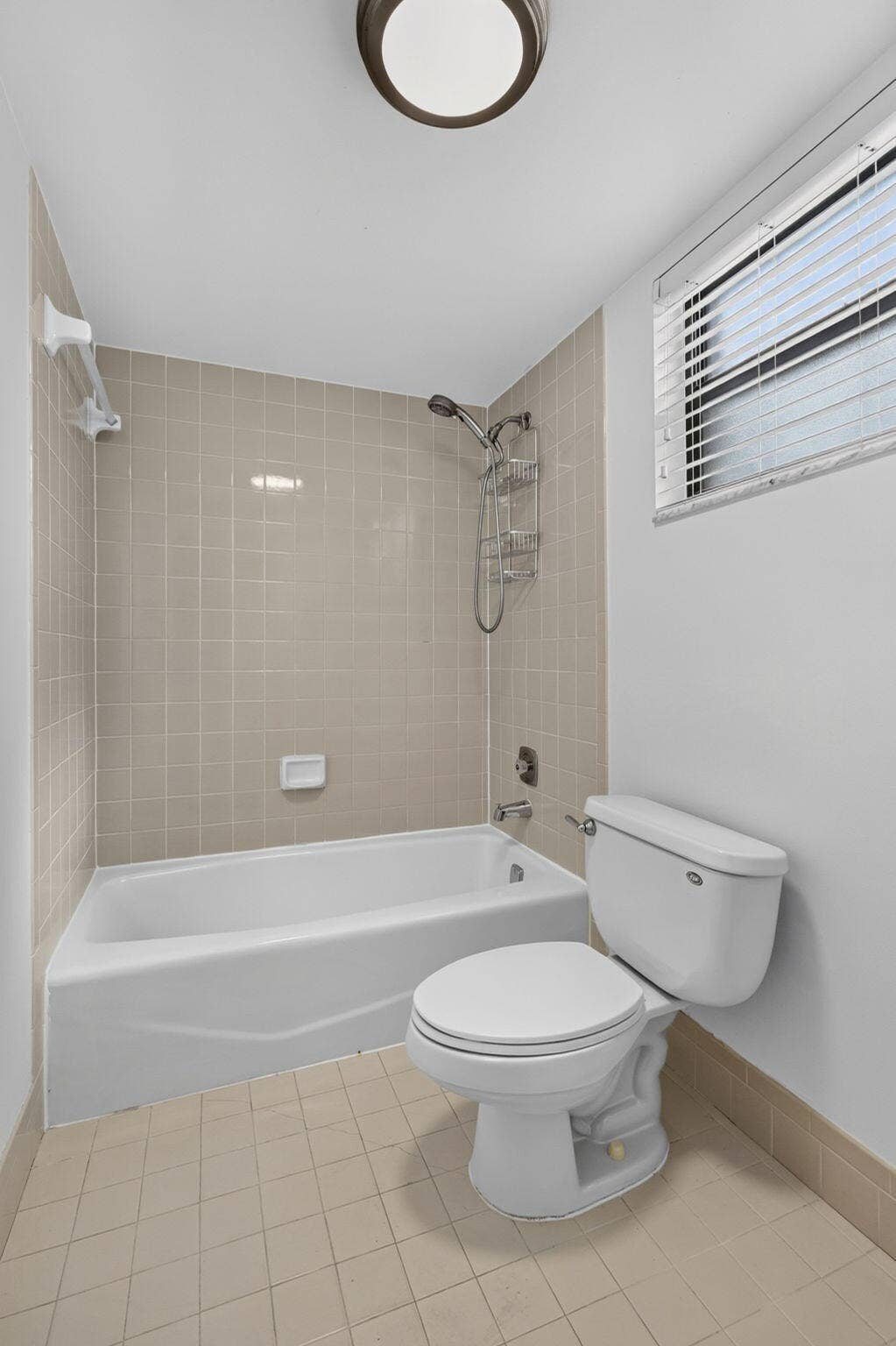Bathroom featuring a built-in bathtub with a tile surround, window with horizontal blinds, flush-mount lighting, and neutral floor tiling