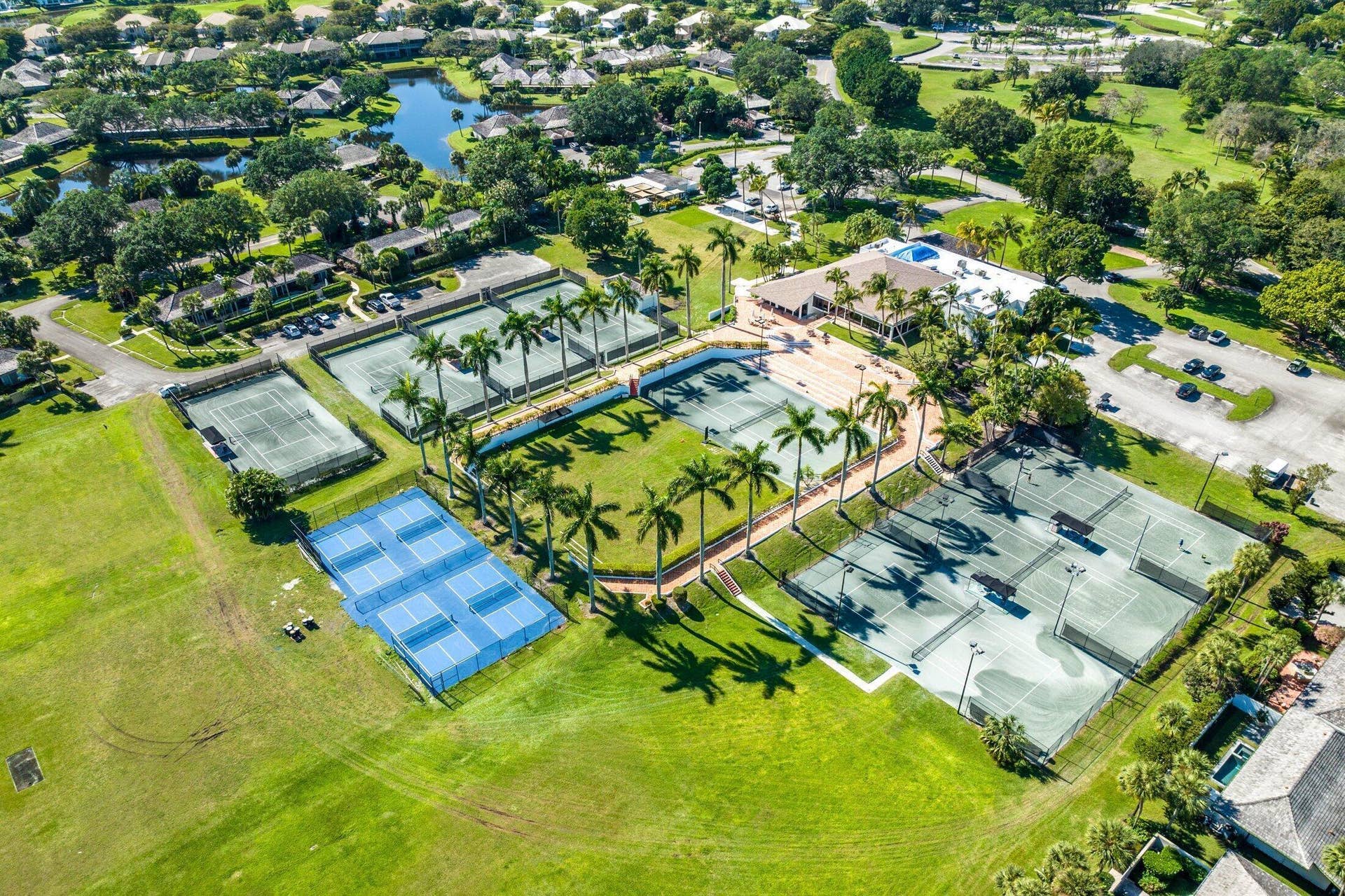 Aerial perspective of a multi-court athletic facility featuring multiple tennis courts, pickleball courts, manicured landscaping, and palm trees