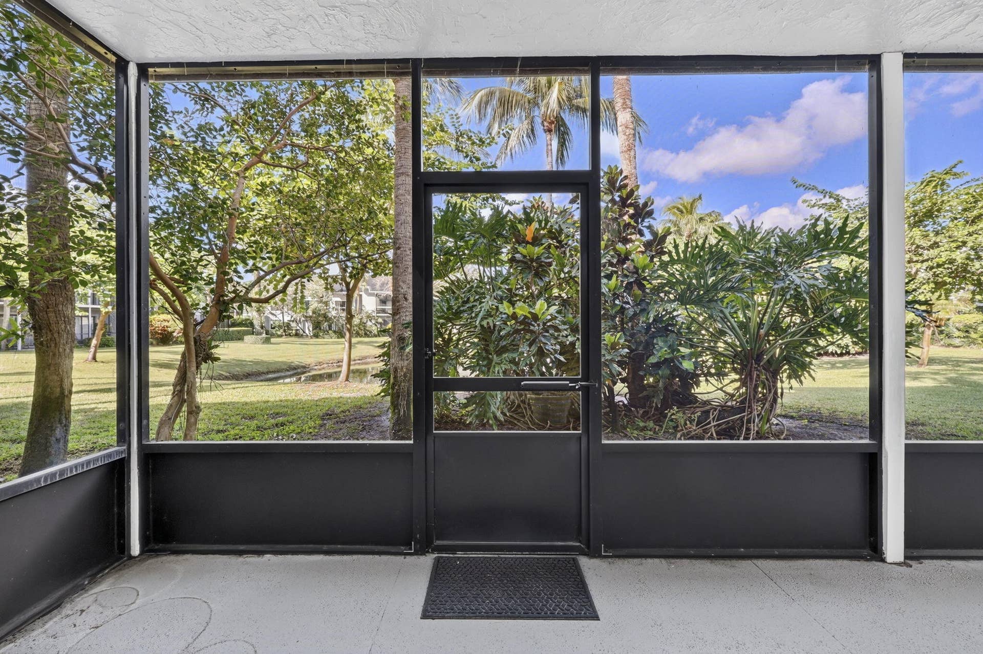 Screened-in porch featuring a black aluminum frame, concrete flooring, and a full-height screen door with access to a landscaped exterior with mature trees