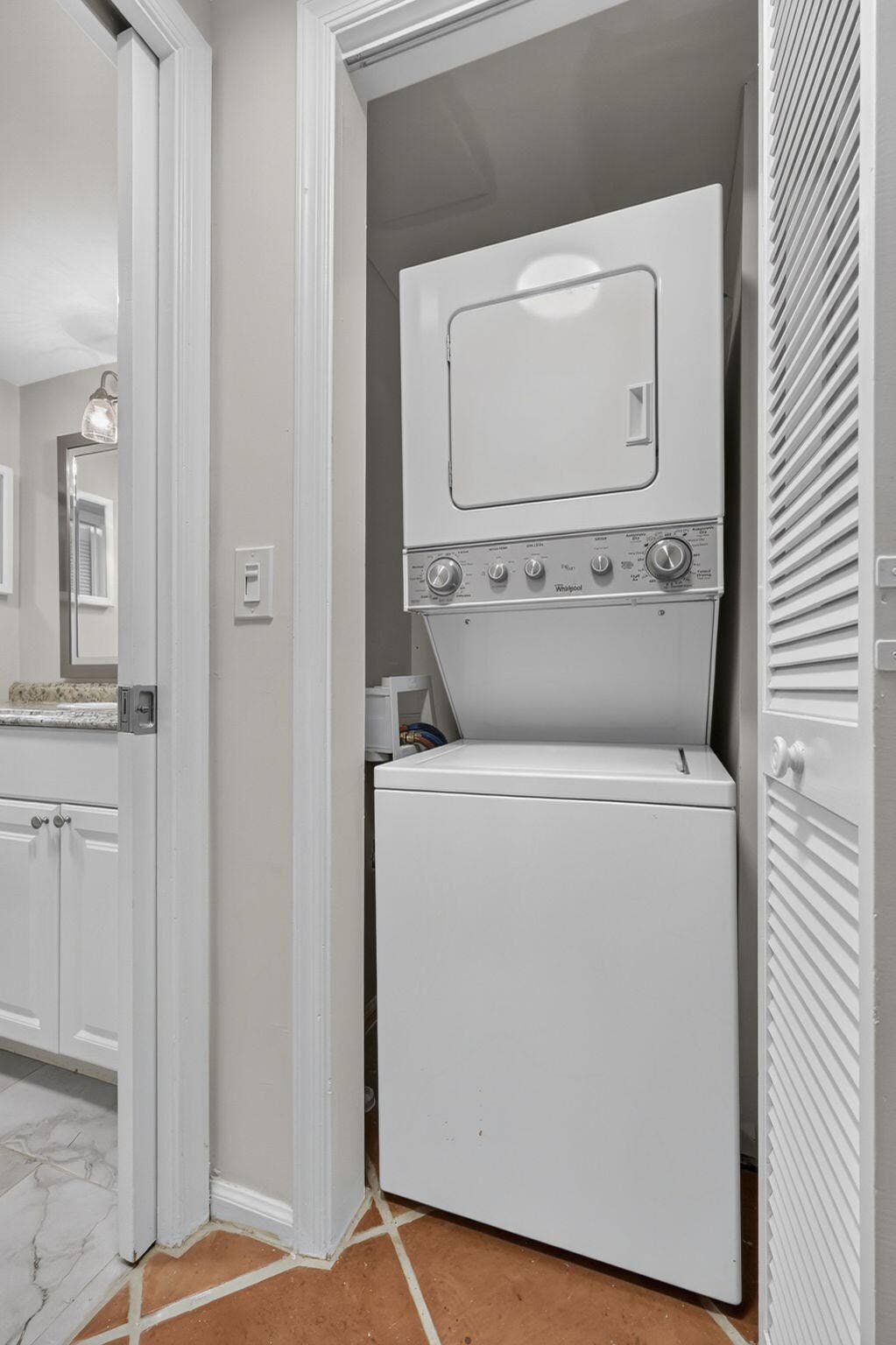 Stacked laundry unit featuring white appliances housed within a closet with a louvered door, complemented by earth-toned tile flooring