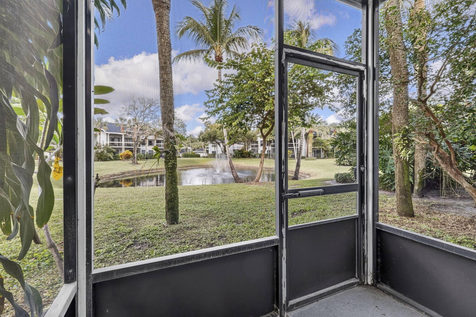 Screened-in enclosure featuring a metal-framed door, concrete flooring, exterior foliage, and a pond with a fountain