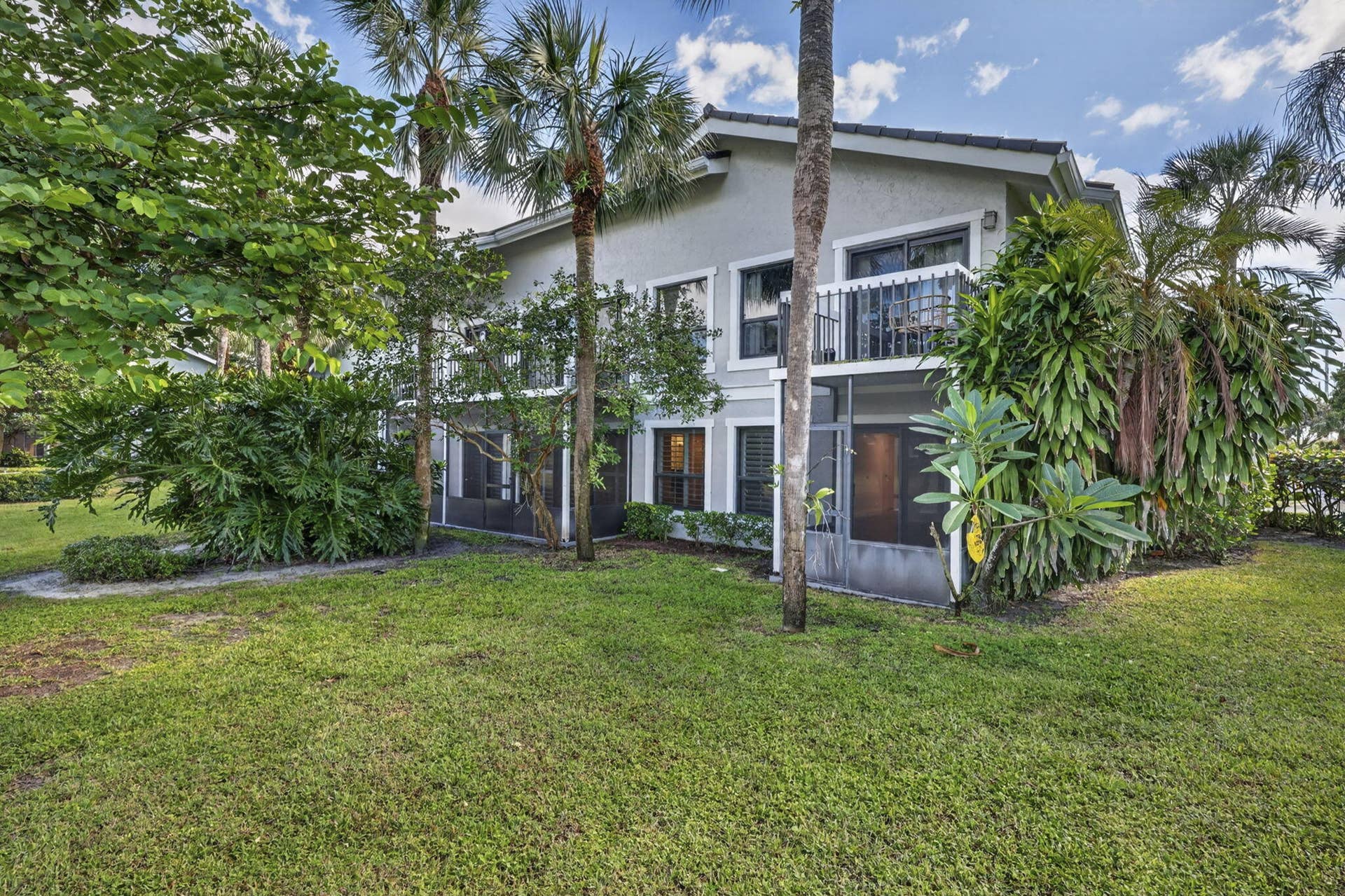 Two-story exterior featuring light-colored stucco siding, a shingled roof, a second-level balcony with metal railing, and screened lower-level enclosures, all set within a mature landscape