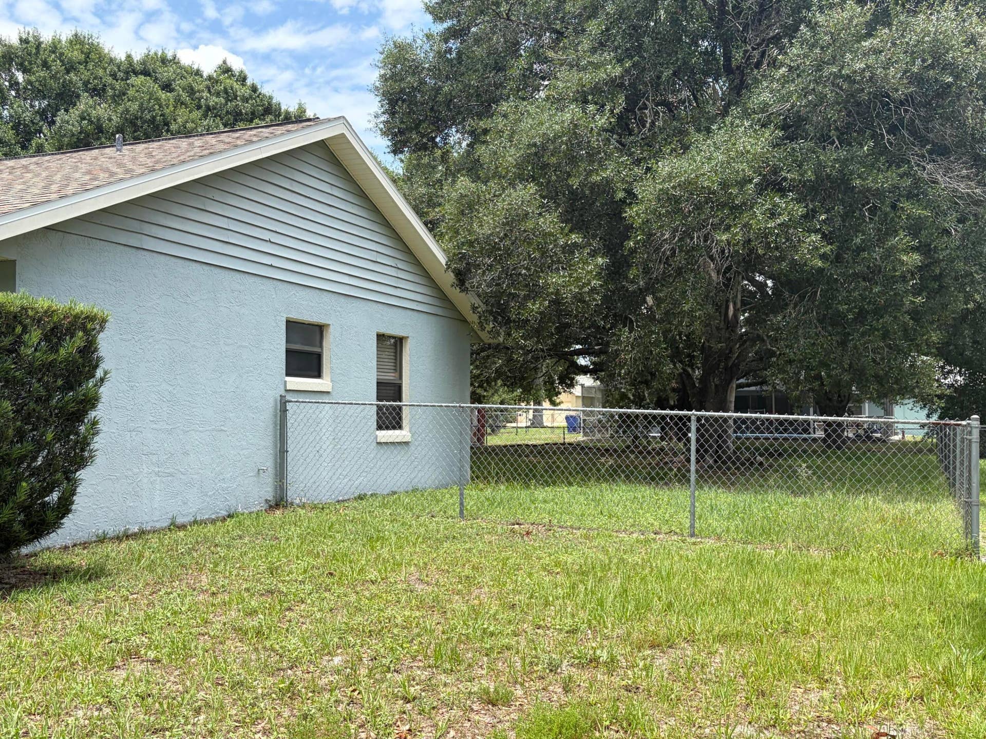 Stucco exterior featuring horizontal siding gable detail, asphalt shingle roofing, and a chain-link fenced backyard with mature trees
