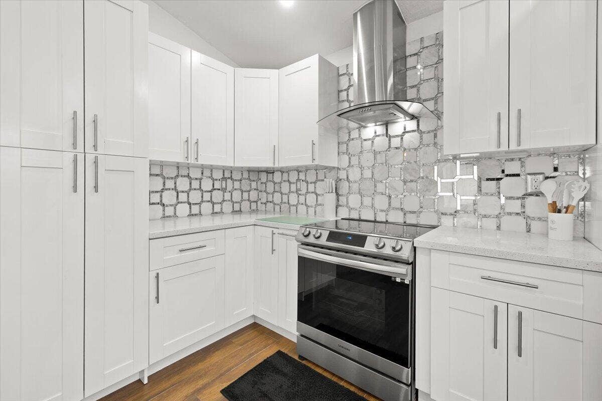 Kitchen featuring white shaker-style cabinetry, stainless steel range with overhead vent hood, light-toned stone-finish countertops, geometric-patterned tile backsplash, and wood-finish flooring