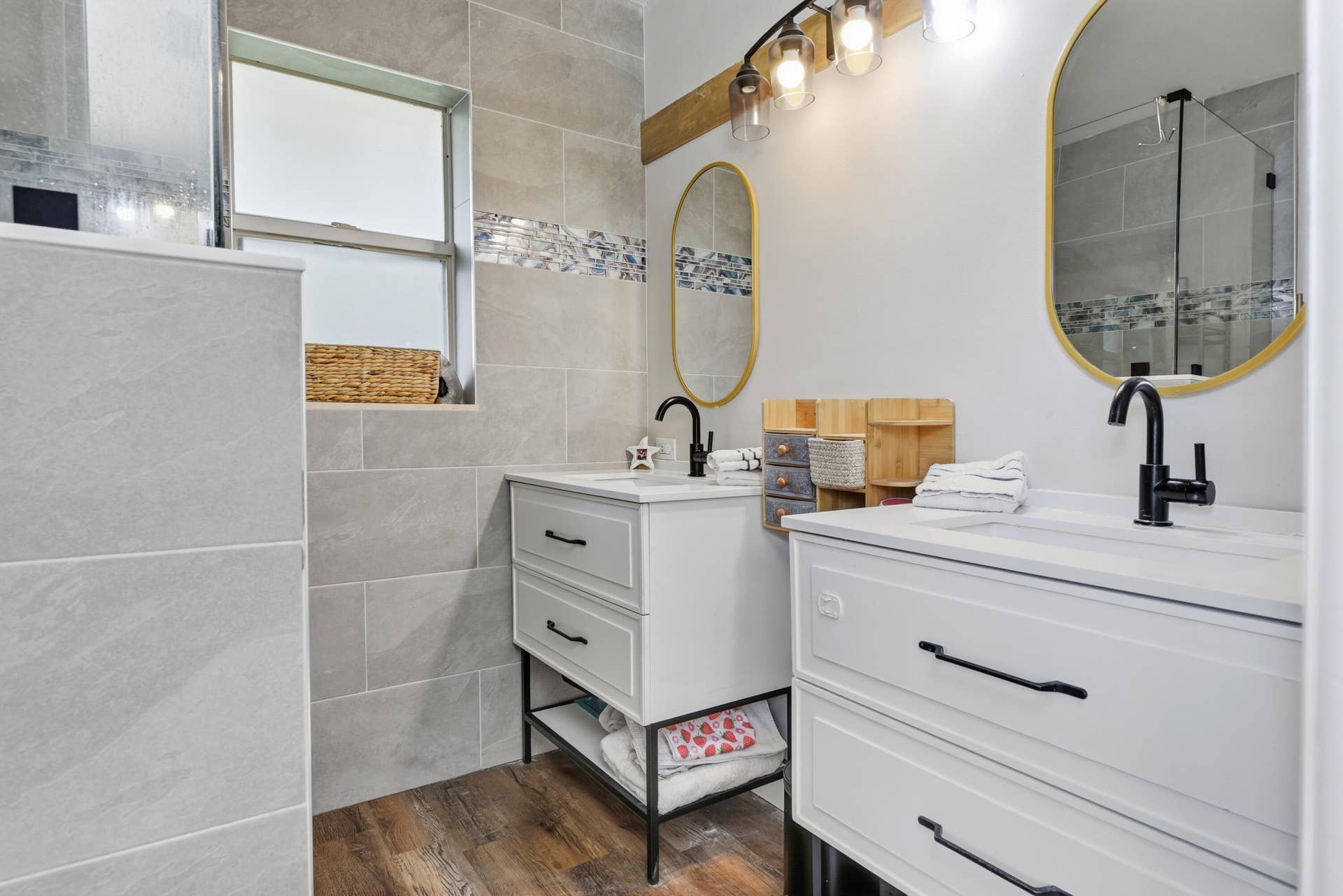 3233 SW SUNSET TRACE Circle, Palm City, FL 34990 Dual vanity with white cabinetry, matte black hardware, integrated rectangular sinks, and wood-finish flooring, complemented by neutral oversized wall tile and arched mirrors with gold-tone frames