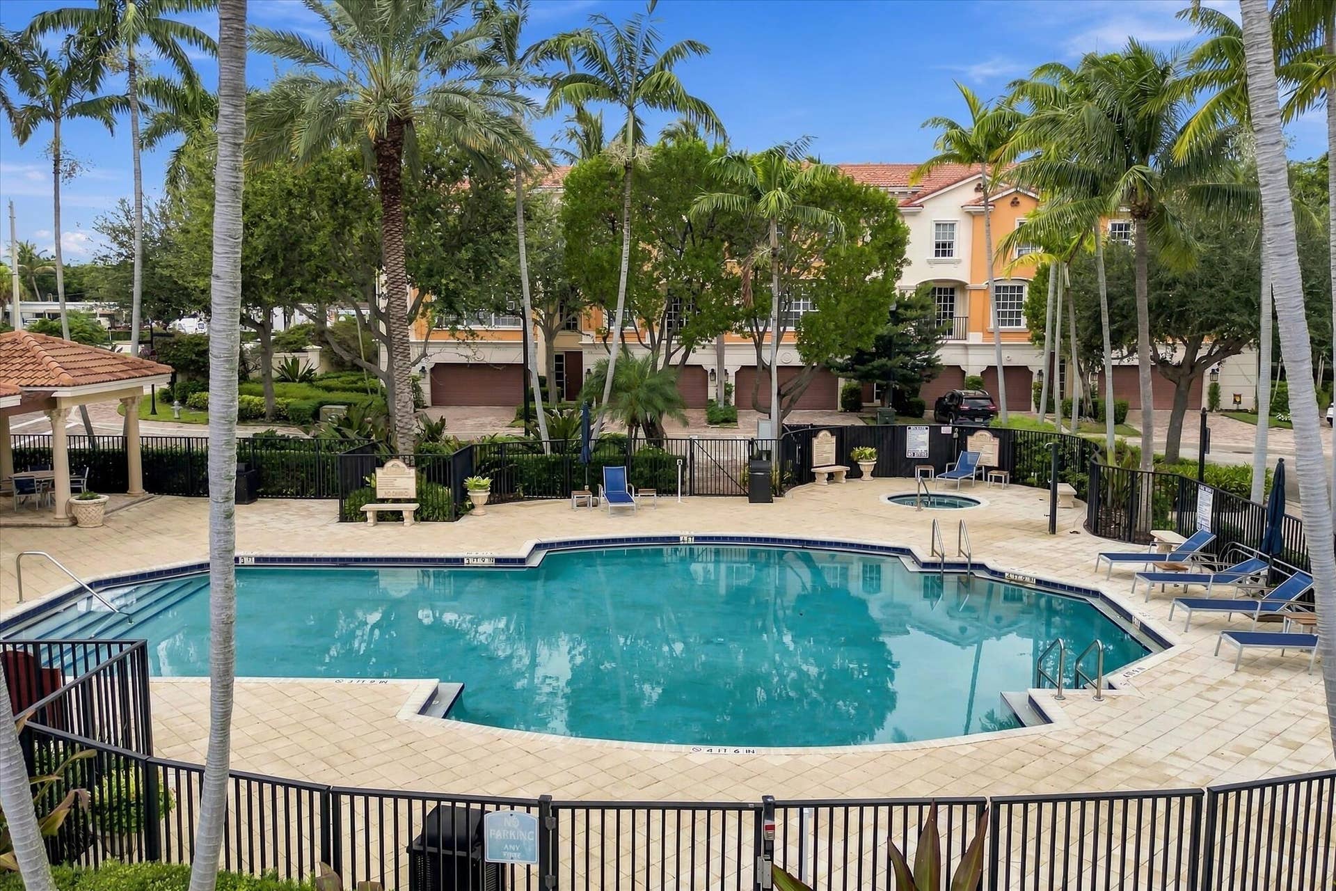 5545 NE Trieste Way, Boca Raton, FL 33487 Swimming pool area featuring stone paver decking, a spa, covered pavilion, and metal perimeter fencing surrounded by mature palm trees