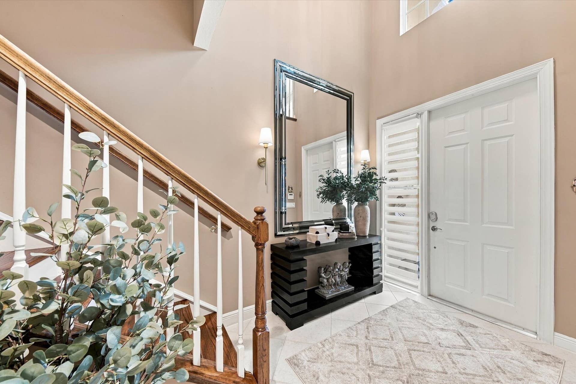 5545 NE Trieste Way, Boca Raton, FL 33487 Foyer featuring a double-height ceiling, staircase with wood handrails and white balusters, multi-panel entry door with sidelight, tile flooring, and wall-mounted light fixtures