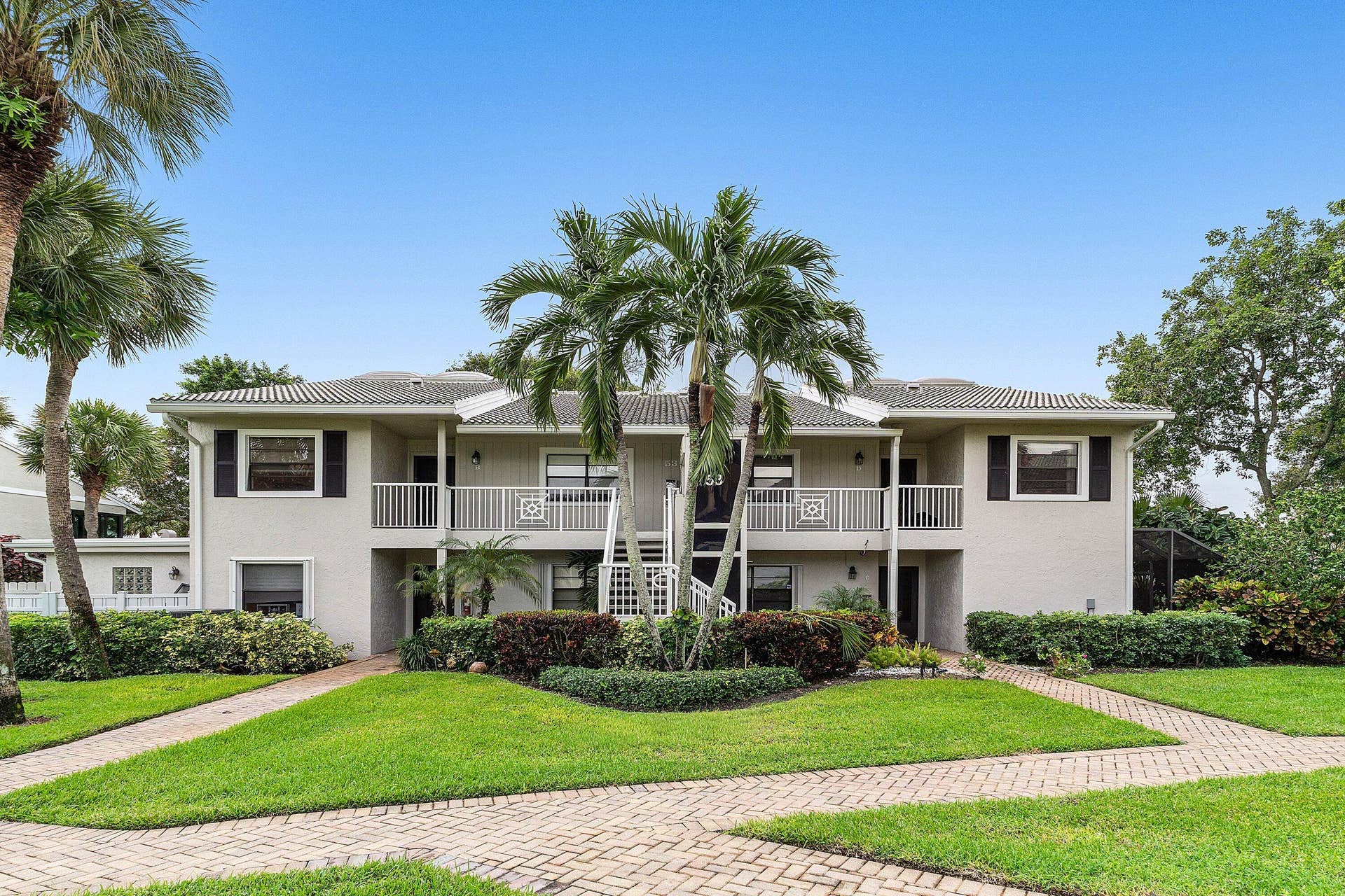 53 EASTGATE Drive, #B, Boynton Beach, FL 33436 Two-story exterior featuring light-toned stucco siding, a tiled roof, upper-level balconies with white railings, and paver walkways surrounding a manicured lawn