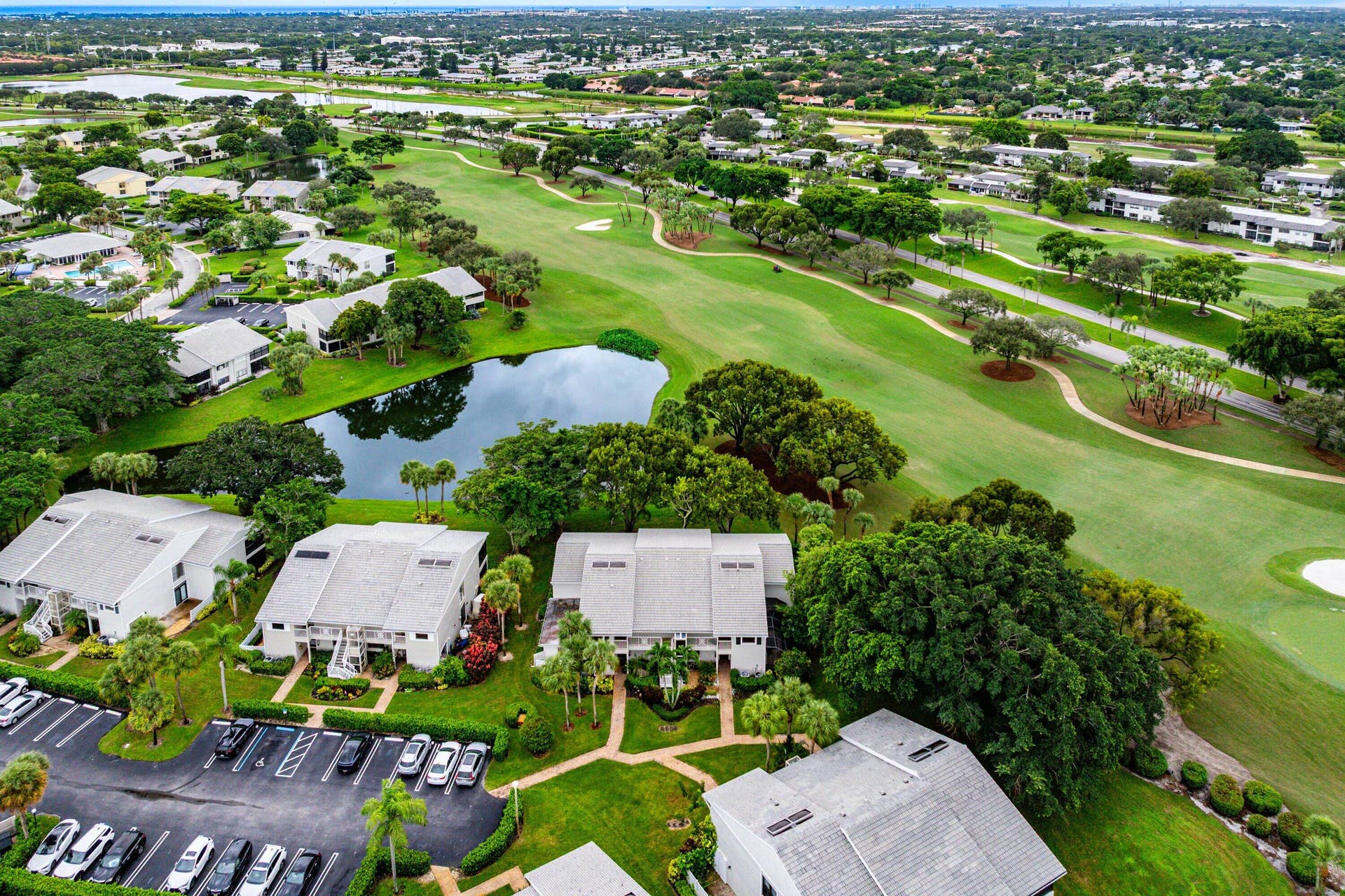 53 EASTGATE Drive, #B, Boynton Beach, FL 33436 Aerial perspective of multi-family residences featuring light-colored shingled roofs, landscaped grounds with mature trees, a central pond, and proximity to golf course fairways