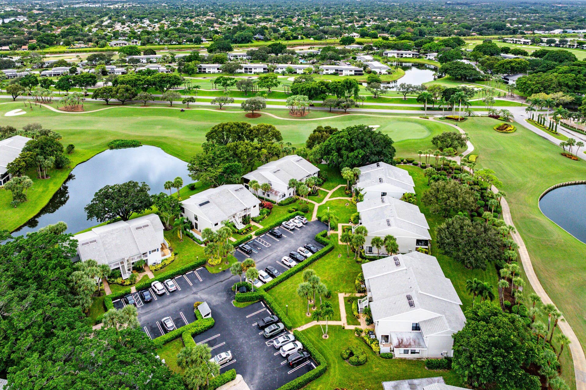 53 EASTGATE Drive, #B, Boynton Beach, FL 33436 Aerial perspective of residential structures featuring light-toned pitched roofs, paved surface parking, manicured grounds, mature trees, and direct proximity to a golf course and water features